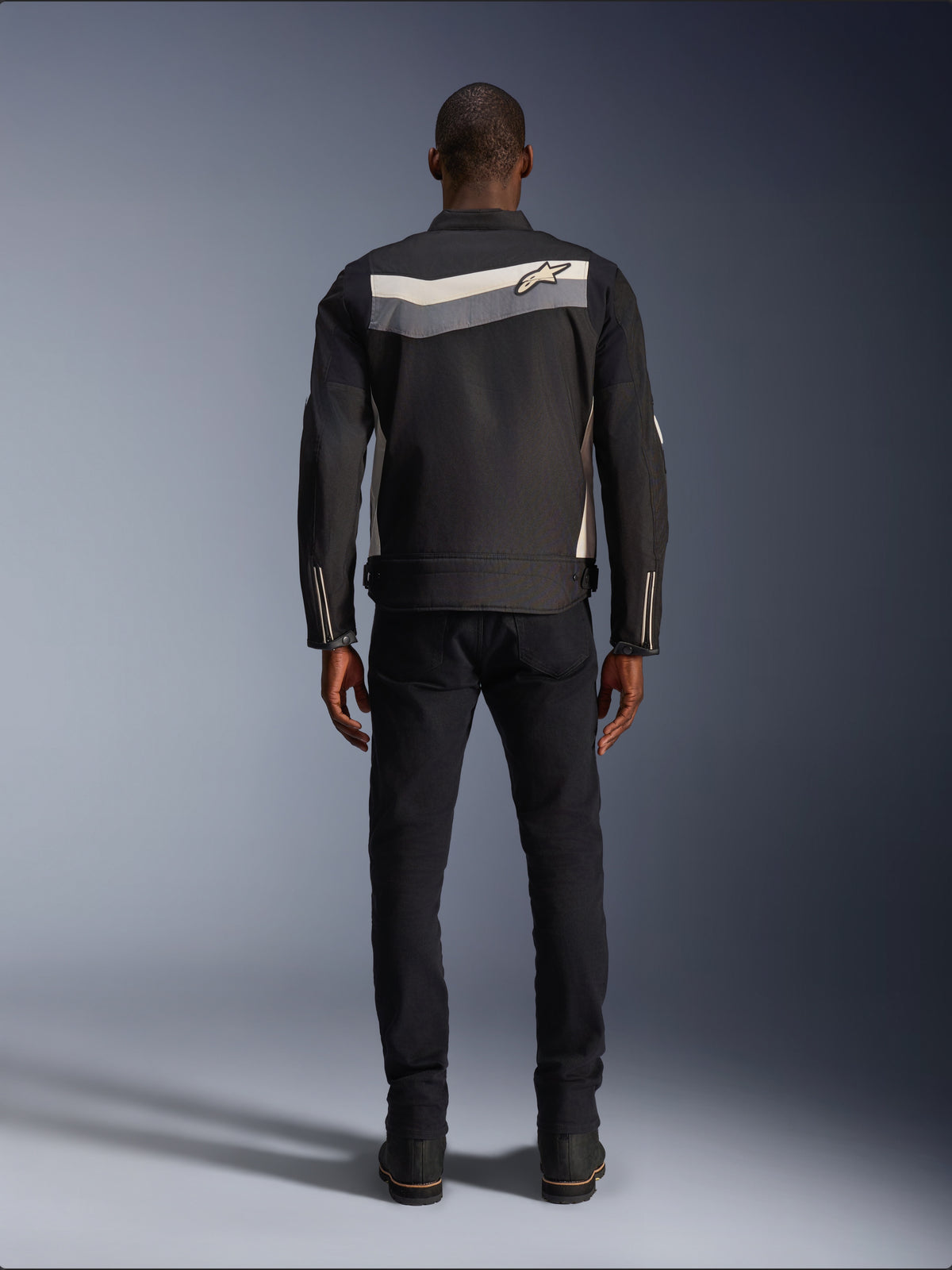 Alpinestars T-Dyno WR Jas, textiele motorjas, zwart, donkergrijs en ecru, achteraanzicht met horizontale streep over de schouders en klein sterlogo, gedragen door model met zwarte broek en laarzen, vintage-geïnspireerde motorkleding