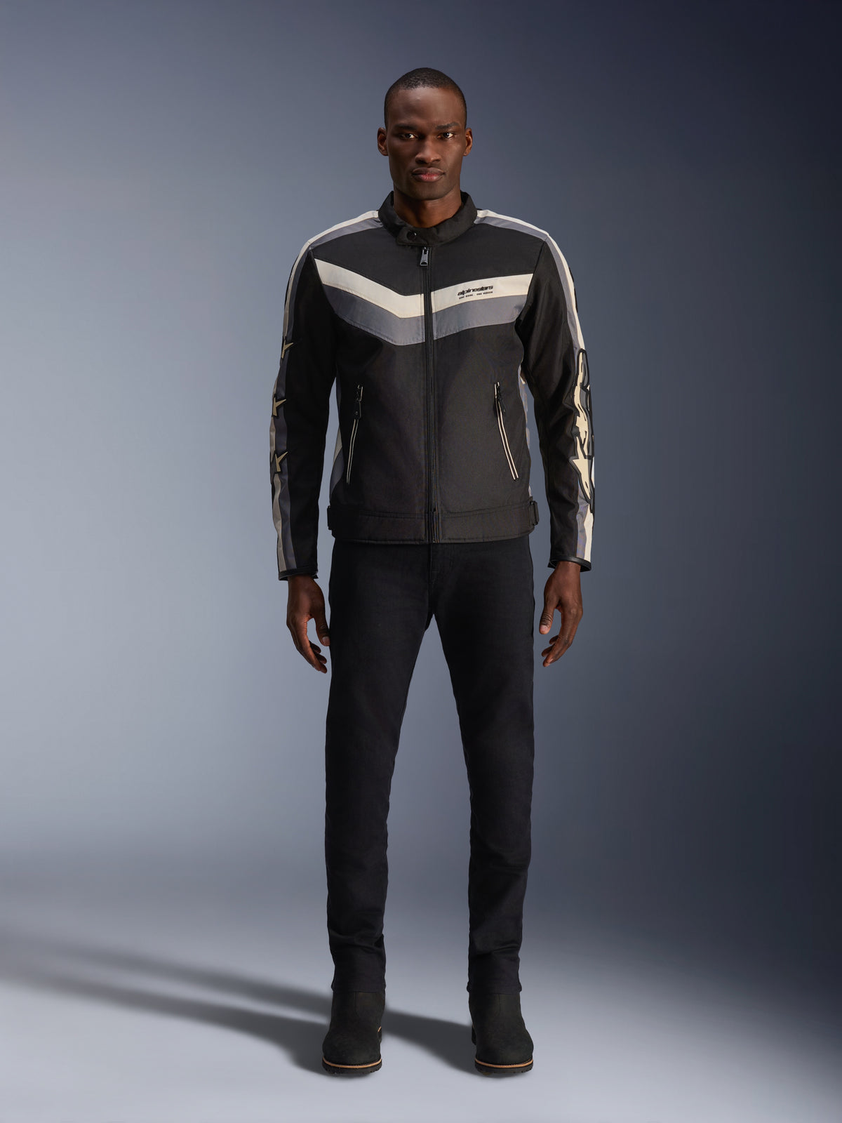 Alpinestars T-Dyno WR Jas, zwart donkergrijs en ecru, textiele motorjas met vintage-geïnspireerd chevronpatroon op de borst, gedragen door een mannelijk model met zwarte motorbroek en laarzen, urban contemporary motorkleding