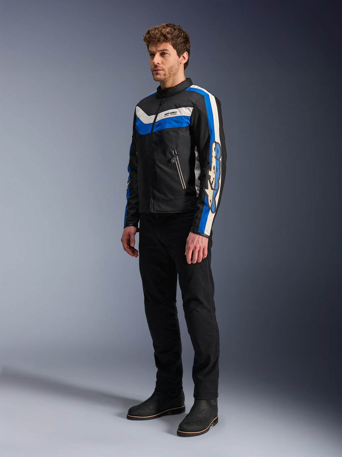 Alpinestars T-Dyno WR Jacke, Schwarz, Blau und Ecru, Textil-Motorradjacke getragen von Model mit schwarzer Hose und Stiefeln, Vintage-inspiriertes Design mit markanten blauen und ecrufarbenen Streifen und Branding an den Ärmeln, Studiofotografie