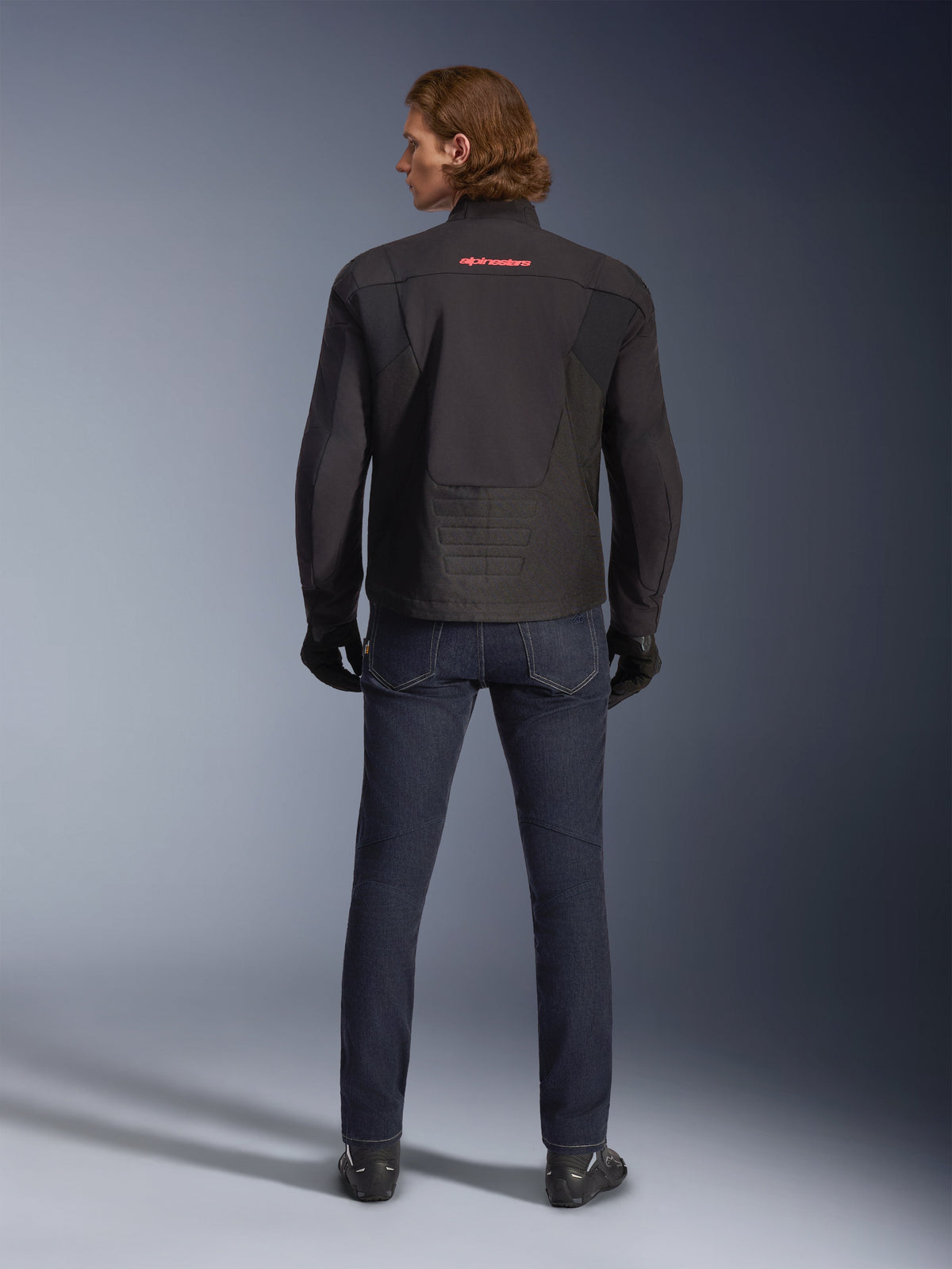 Alpinestars SMX Waterproof Jacket, zwart, achteraanzicht gedragen door een model, met een rood logo op de bovenrug, verlengde achterzoom voor dekking tijdens het rijden, gecombineerd met donkerblauwe motorjeans en zwarte handschoenen.