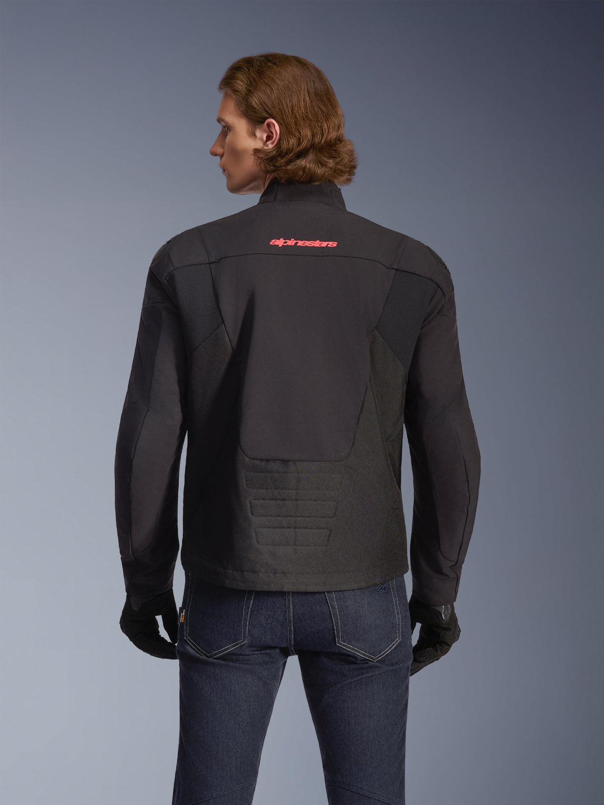 Alpinestars SMX Waterproof Jas, zwart met felrood logo, achteraanzicht getoond op een model, gedragen met donkerblauwe jeans en zwarte handschoenen, textiele motorjas voor weg- en sportief rijden