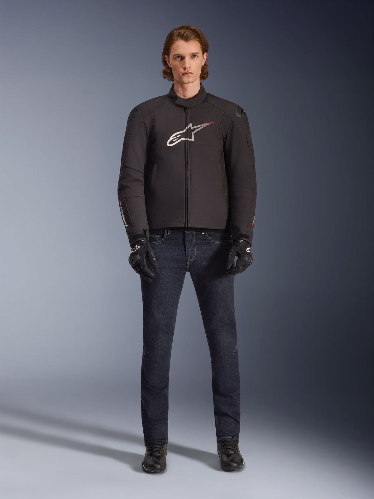 Alpinestars SMX Waterproof Jacket in zwart met wit en rood logo, model draagt donkere denim motorbroek, zwarte handschoenen en motorlaarzen, sportieve motoroutfit voor alle weersomstandigheden