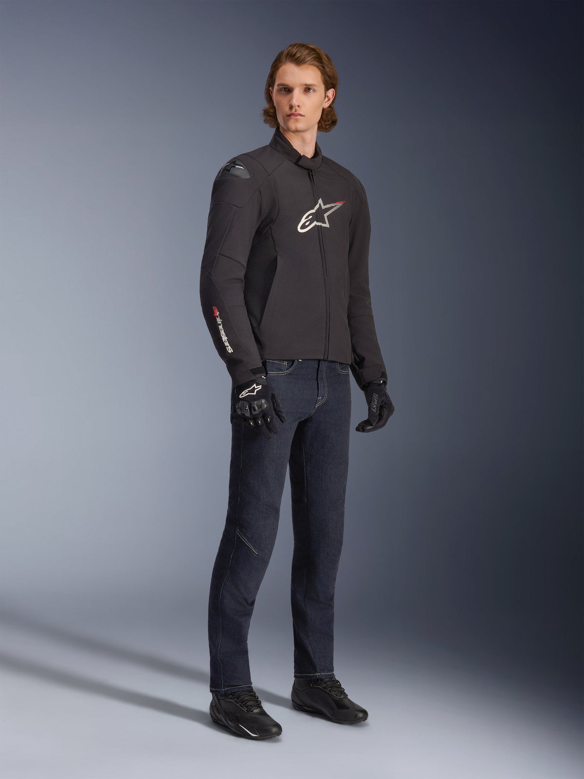Mannelijk model draagt Alpinestars SMX Waterproof Jacket, zwart met witte en rode logo-accenten, gecombineerd met een donkerblauwe denim broek en zwarte motorhandschoenen, strakke sport-fit textiel motorjas