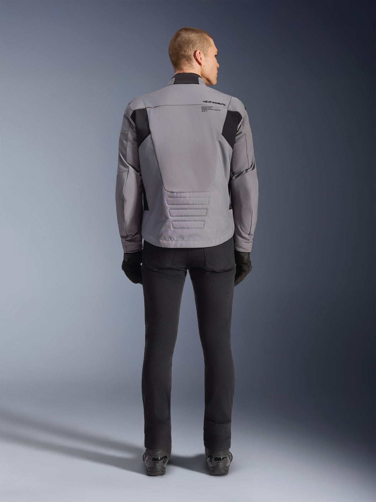Alpinestars SMX Waterproof Jacket, achteraanzicht, donkergrijs en zwart, voorzien van zwarte textielpanelen op de schouders en een sportieve pasvorm, gecombineerd met een zwarte broek en handschoenen voor een complete motorlook