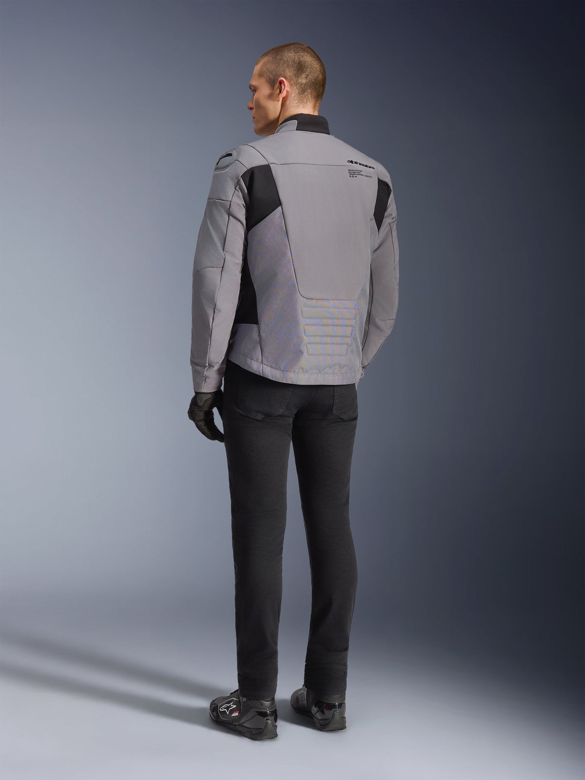Alpinestars SMX Waterproof Jacket in donkergrijs en zwart, achteraanzicht op een model, gedragen met een zwarte broek en laarzen, voorzien van een technische textielconstructie met zwarte panelen en Alpinestars-branding op de schouder.