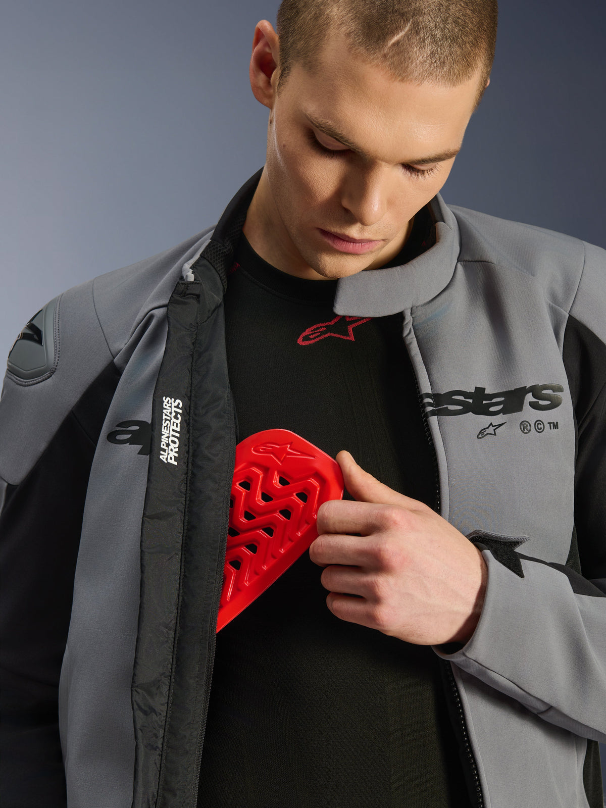 Mannelijk model draagt de Alpinestars SMX Waterproof Jacket in Dark Gray Black en plaatst een rode Nucleon borstprotector in een binnenzak, waarbij de compatibiliteit voor protectie en de beschermende kenmerken van de jas worden getoond.