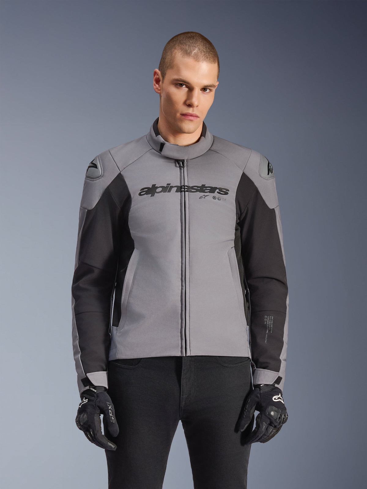 Alpinestars SMX Waterproof Jacket, sportieve motorjas, donkergrijs en zwart, met een hoge kraag en DFS Lite schoudersliders, gedragen met zwarte SMX-handschoenen, strakke atletische pasvorm voor rijden in alle weersomstandigheden