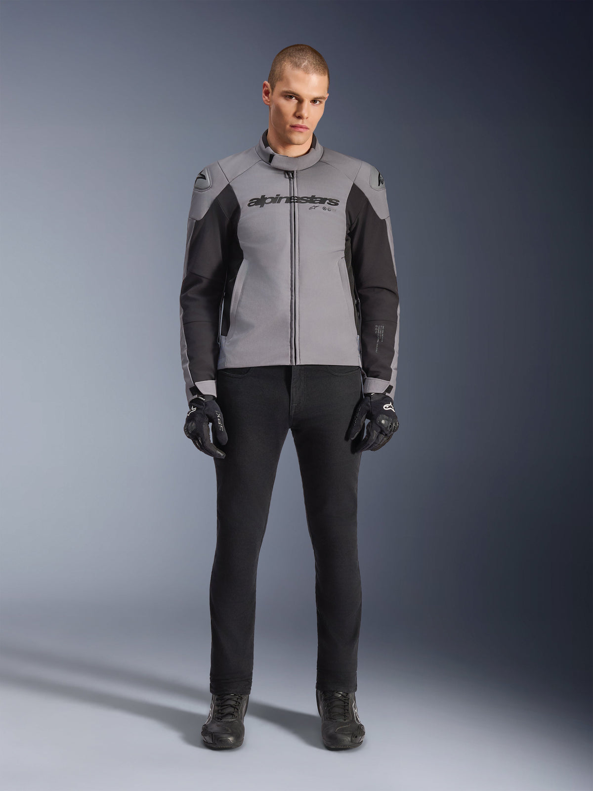 Alpinestars SMX Waterproof Jas, donkergrijs en zwart, vooraanzicht gedragen door een mannelijk model, textiele sportieve motorjas met zwarte zijpanelen en Alpinestars-branding op de borst, gecombineerd met een zwarte motorbroek en handschoenen.