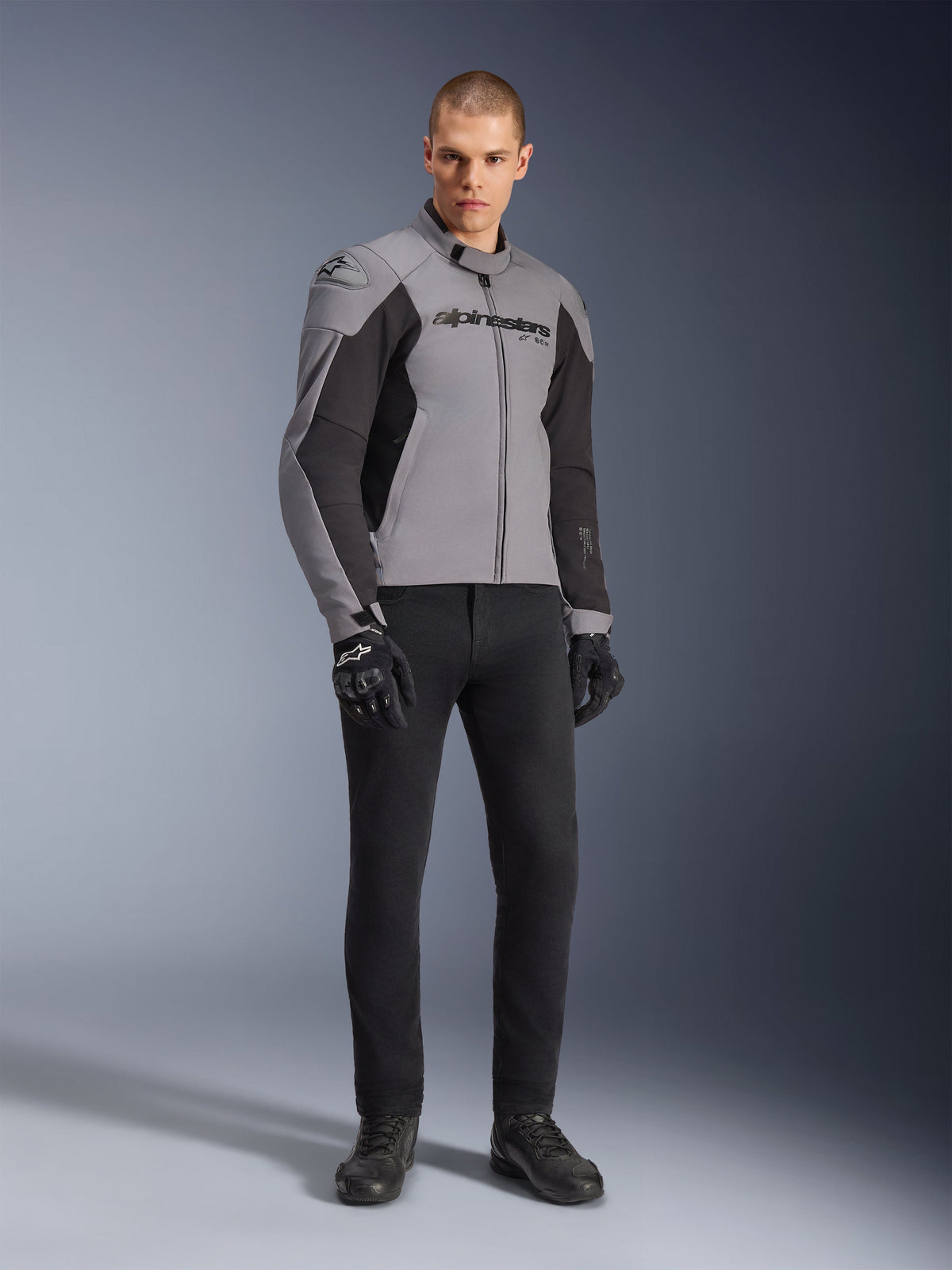 Alpinestars SMX Waterproof Jacket, motorjas, donkergrijs en zwart, gedragen door model met zwarte motorhandschoenen en zwarte broek, met opvallend logo op de borst en een technisch sport-mapped ontwerp voor motorrijden onder alle weersomstandigheden.