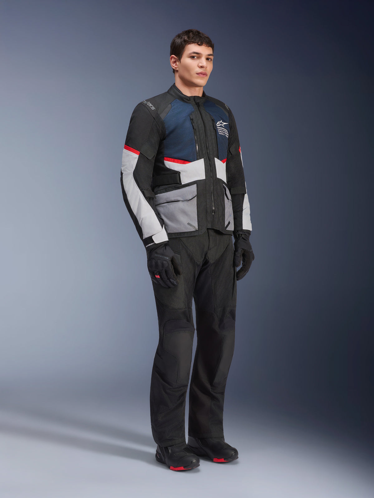 Mannelijk model draagt de Alpinestars Andes Air Drystar® Jas in Deep Blue Black Ice Gray, gecombineerd met een zwarte motorbroek, handschoenen en laarzen, voorzien van grote mesh-panelen voor ventilatie, lichtgrijze zakken en rode accenten, ontworpen voor adventure touring.