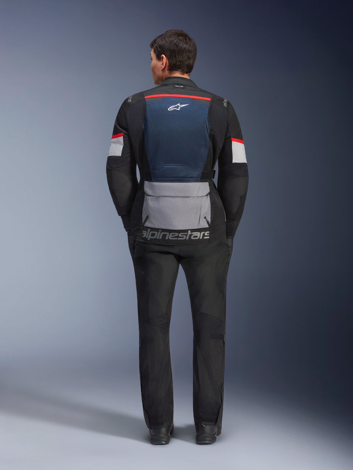 Alpinestars Andes Air Drystar jas, Deep Blue Black Ice Gray, achteraanzicht op model, voorzien van een blauw mesh rugpaneel voor ventilatie, een grote grijze zak onderaan de rug, rode en witte kleuraccenten op de mouwen en het 'alpinestars' woordmerk, gecombineerd met een zwarte motorbroek voor een complete touringoutfit.