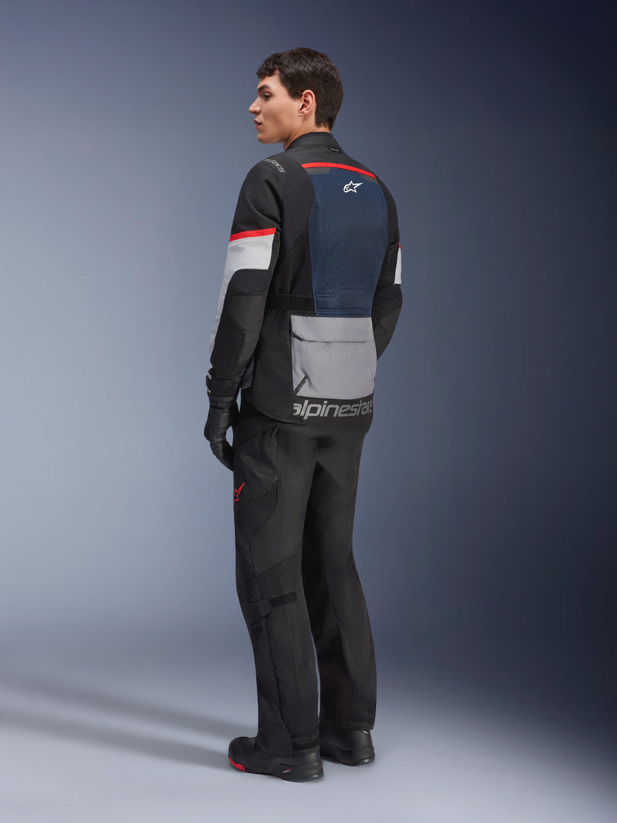 Mannelijk model van achteren en opzij gezien, dragend de Alpinestars Andes Air Drystar® Jas in Deep Blue Black Ice Gray, gecombineerd met een zwarte motorbroek en handschoenen, voorzien van een blauw mesh achterpaneel, grijze opbergzak op de onderrug en rode accenten op de mouwen, professionele adventure touring uitrusting.