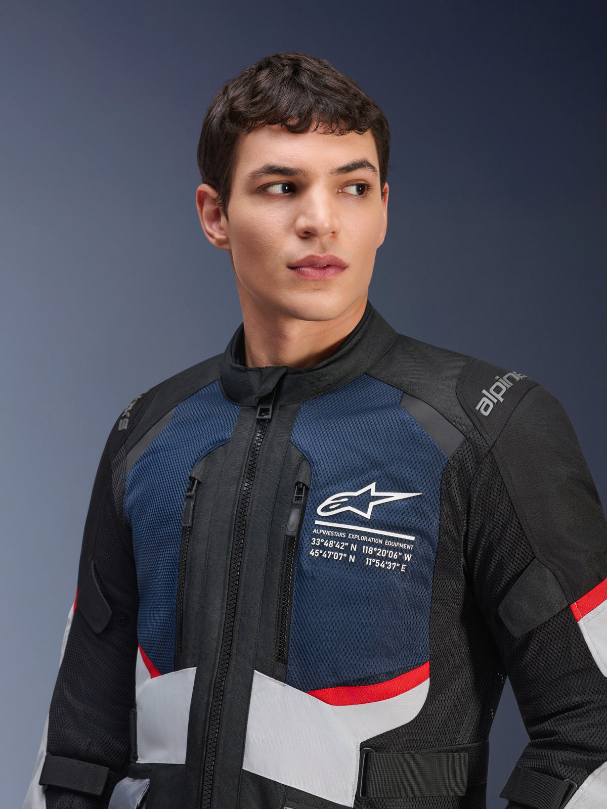 Alpinestars Andes Air Drystar® Jas, motorjas, diepblauw, zwart en ijsgrijs met rode accenten, gedragen door een mannelijk model, voorzien van geventileerde mesh borstpanelen, wit sterlogo met exploratiecoördinaten en weggericht adventure-ontwerp