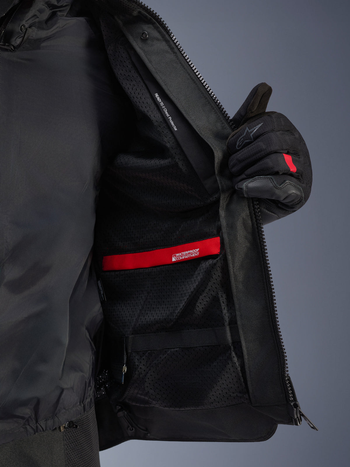 Interieur detailweergave van de Alpinestars Andes Air Drystar® jas, met de ademende zwarte mesh voering, rood waterdicht compartiment en borstprotectorzak, opengehouden door een zwarte motorhandschoen.