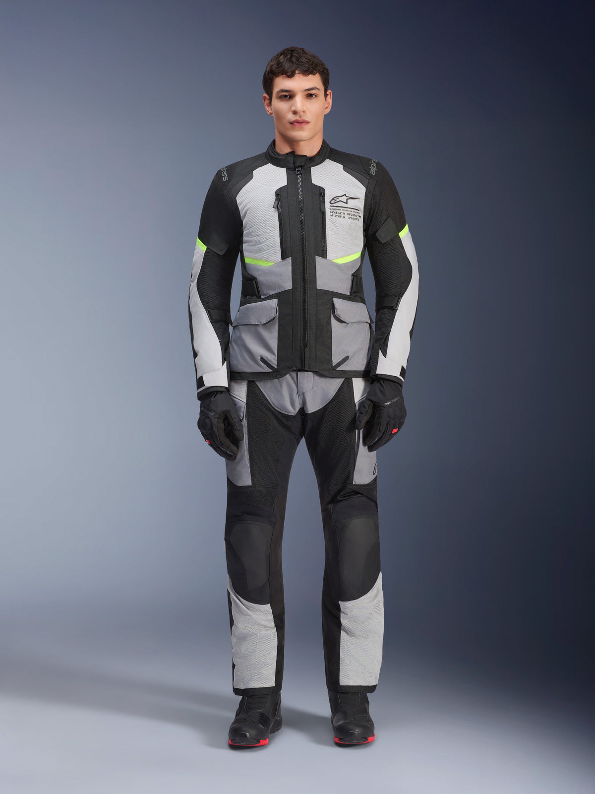 Veste Alpinestars Andes Air Drystar® en Gris Glacé, Gris Foncé et Noir, portée par un modèle masculin avec pantalon et gants de moto assortis, design touring aventure avec de grandes poches avant et des accents jaune fluo, vue de face en studio