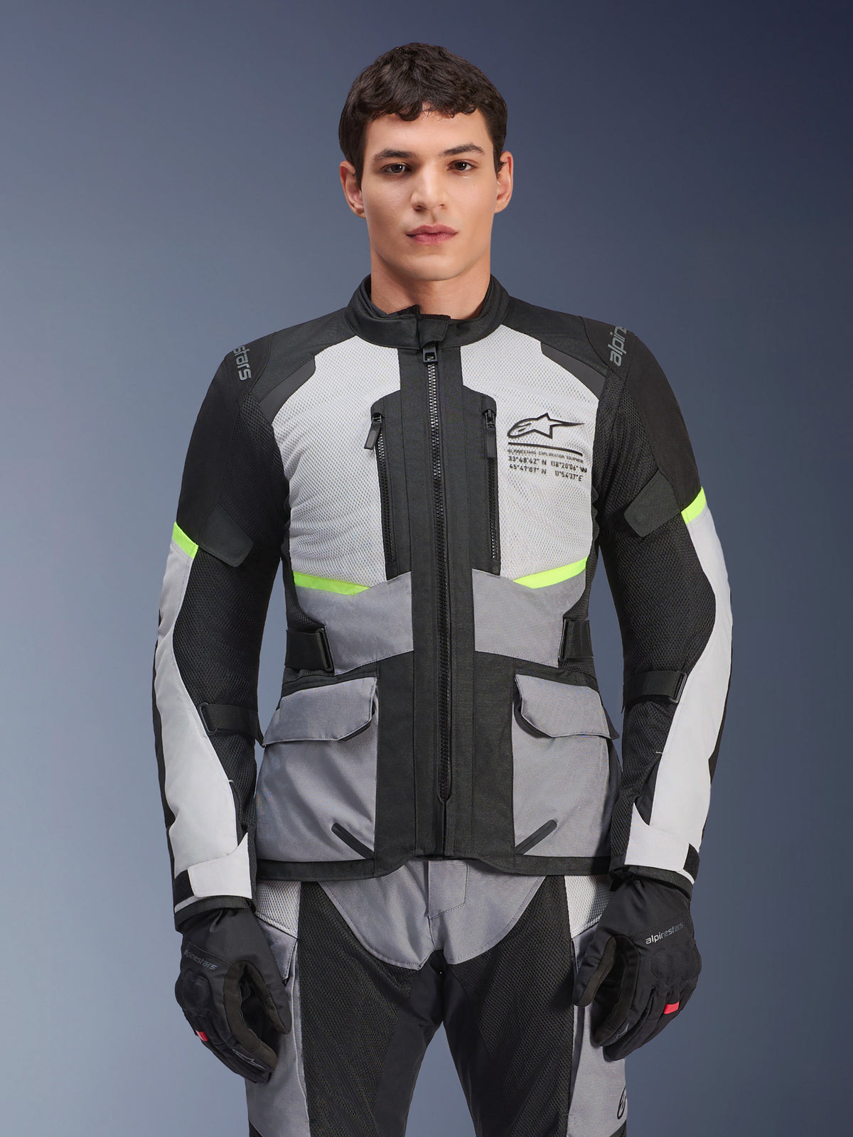 Motard portant la veste Alpinestars Andes Air Drystar® en gris glacier, gris foncé et noir, avec des accents jaune fluo et des panneaux de ventilation en mesh, accompagnée d'un pantalon et de gants de moto assortis, équipement de touring aventure