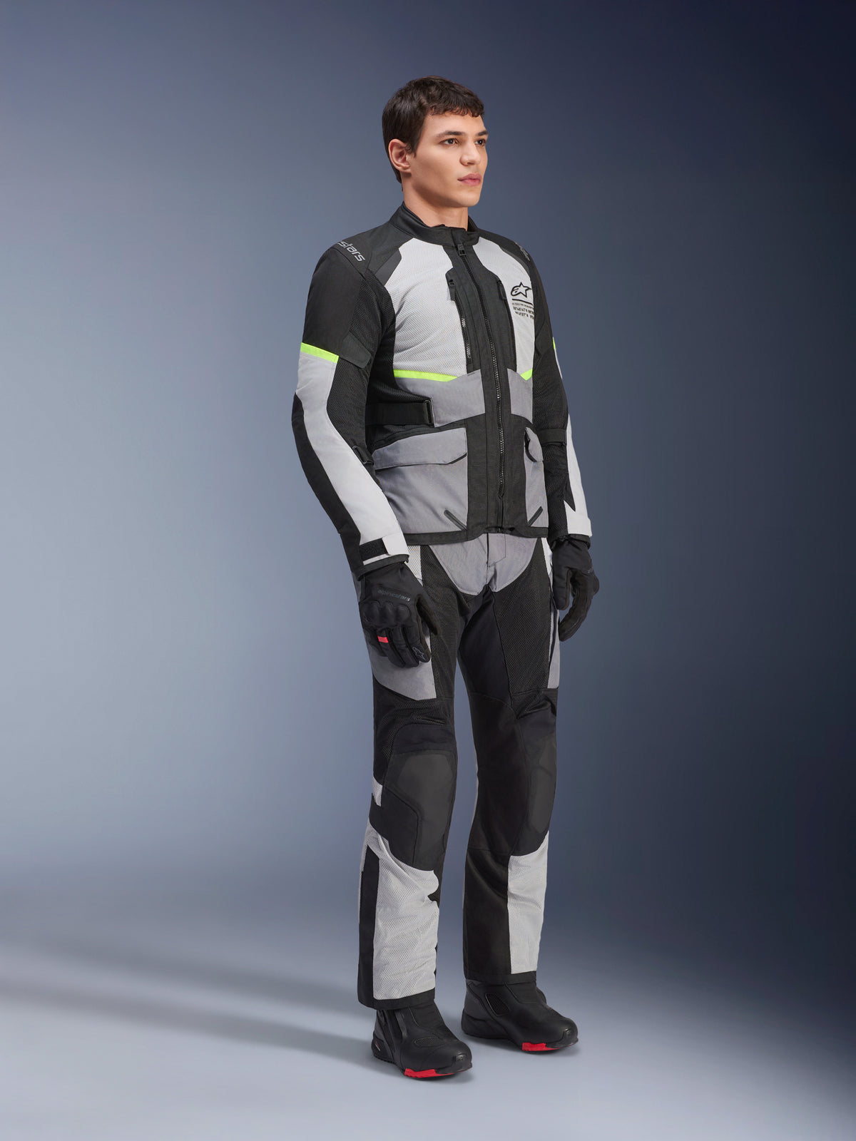 Veste Alpinestars Andes Air Drystar®, gris glacé, gris foncé et noir, portée par un mannequin avec une tenue de touring aventure complète, pantalon assorti et bottes noires, comprenant une ventilation en mesh, des accents néon et le marquage Alpinestars.