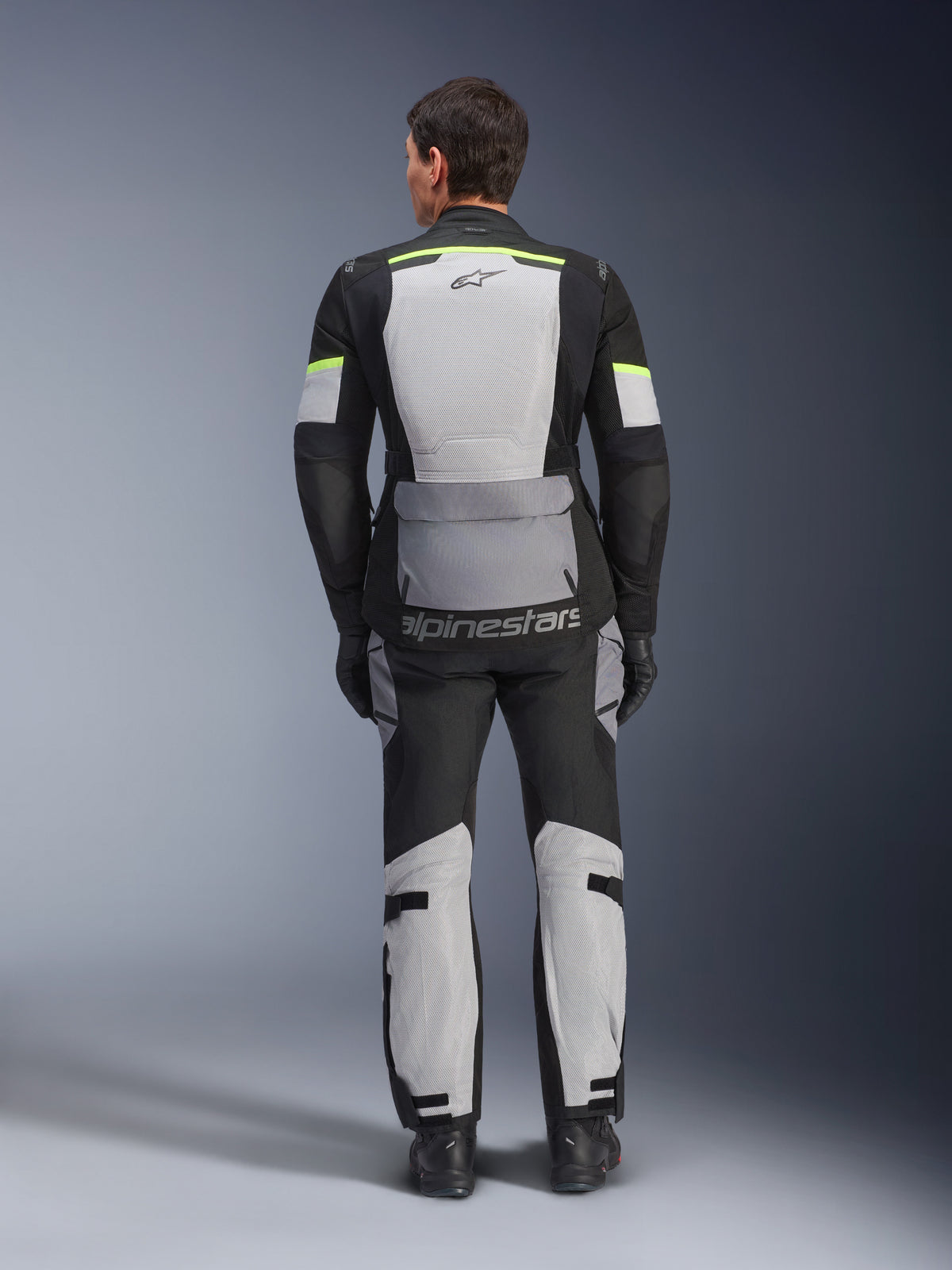 Veste Alpinestars Andes Air Drystar®, veste de touring moto, gris glacé, gris foncé et noir, vue de dos montrant de larges panneaux en mesh pour la ventilation, des accents jaune fluo et le logo Alpinestars, portée avec un pantalon de touring assorti