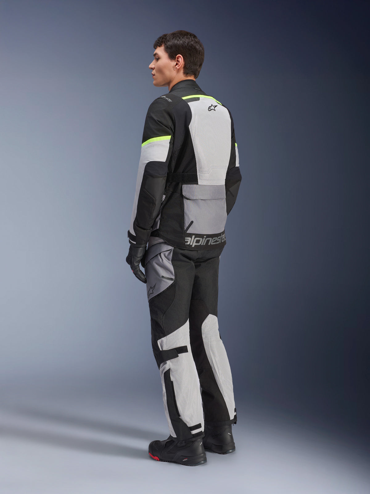 Veste Alpinestars Andes Air Drystar®, veste de moto touring, gris glacé et gris foncé avec empiècements noirs et accents jaune fluo, portée avec pantalon et gants assortis, vue arrière de trois-quarts montrant la ventilation en mesh et le marquage Alpinestars.