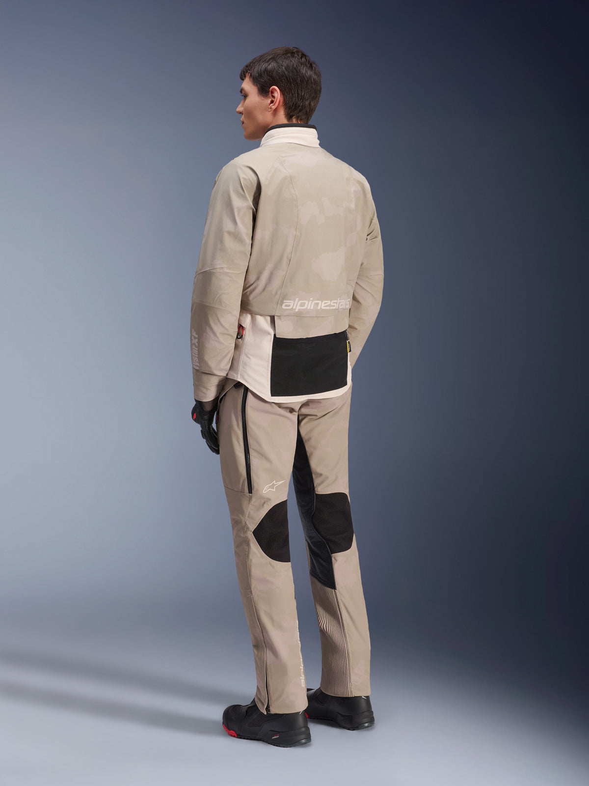 Modell in der Rückansicht mit der AMT-10LAB Drystar® XF Jacke und passender Hose, White Sand Aluminum Camo, Adventure-Motorradausrüstung, mit schwarzen Funktionseinsätzen und Alpinestars-Branding, getragen mit schwarzen Motorradstiefeln und Handschuhen, koordiniertes Outfit für Allwetter-Touren