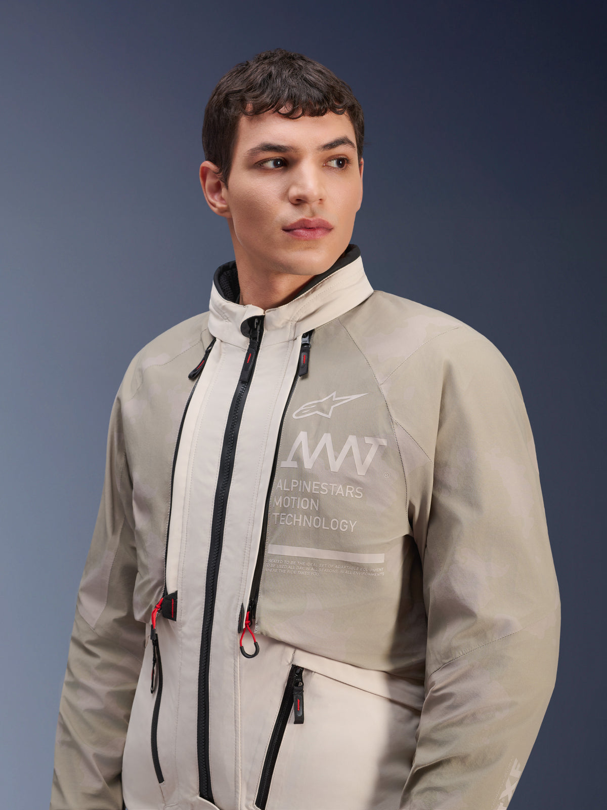 Alpinestars AMT-10LAB Drystar® XF Jacke, White Sand Aluminum Camo, modulare Adventure-Motorradjacke, mit „Alpinestars Motion Technology“-Branding auf der Brust, getragen von einem männlichen Model.