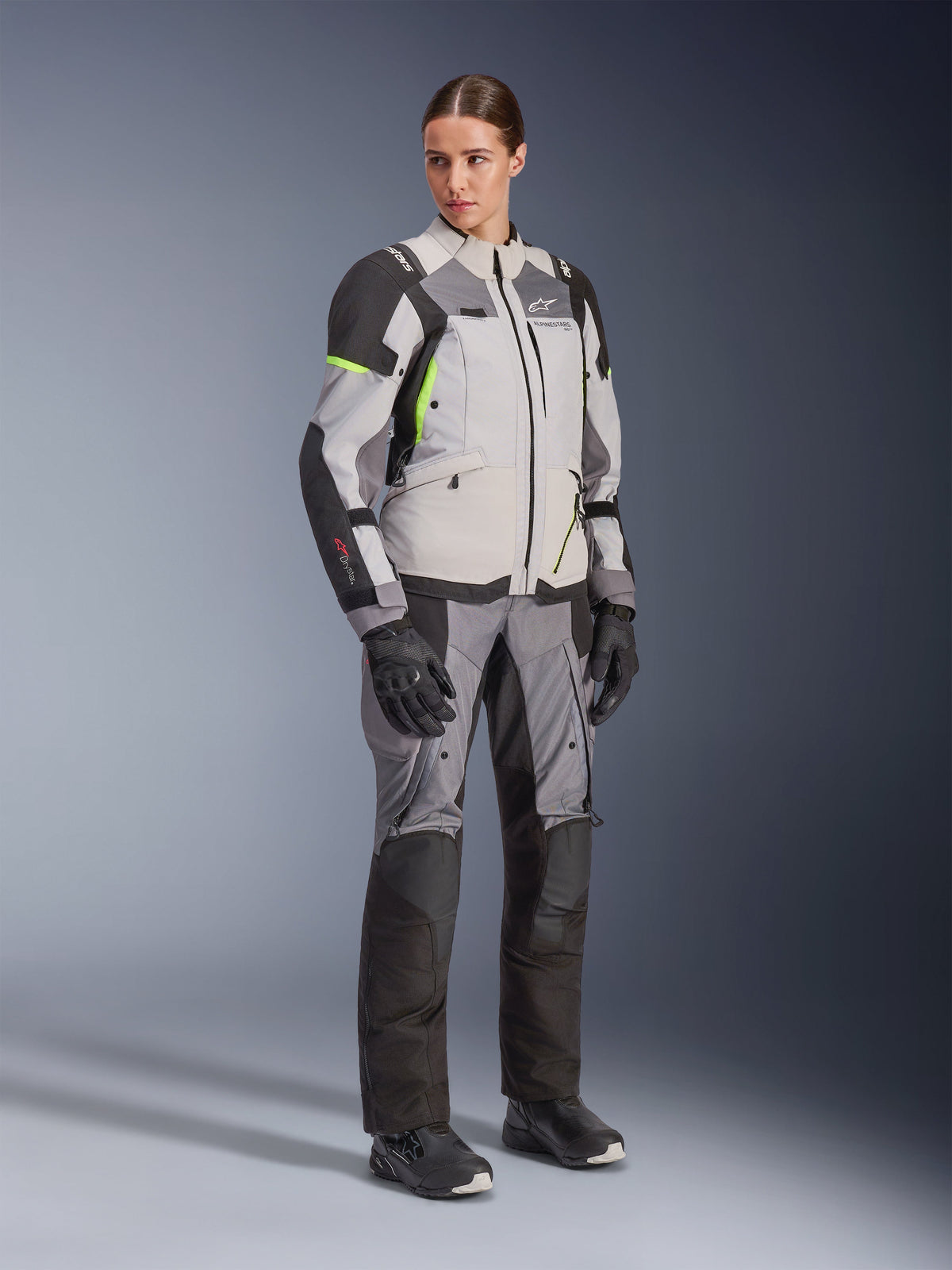 Frau trägt Alpinestars Stella Andes V4 Drystar® Jacke und passende Hose, Ice Gray und Dark Gray mit neongelben Akzenten, komplettes Adventure-Touring-Bekleidungsset mit schwarzen Handschuhen und Stiefeln, Ganzkörper-Studioaufnahme.