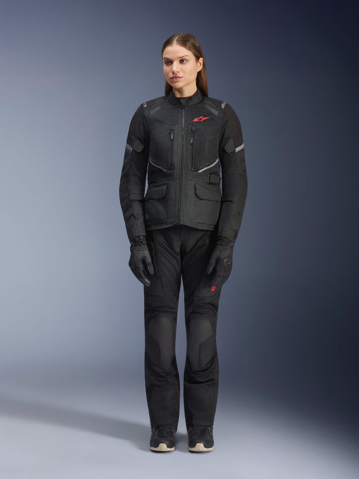 Modèle féminin portant la veste Alpinestars Stella Andes Air Drystar® noire, assortie d'un pantalon de touring et de gants noirs, avec de larges panneaux de ventilation en maille, des touches de logo rouge et des détails réfléchissants, photo de studio professionnelle pour équipement de moto pour femme.