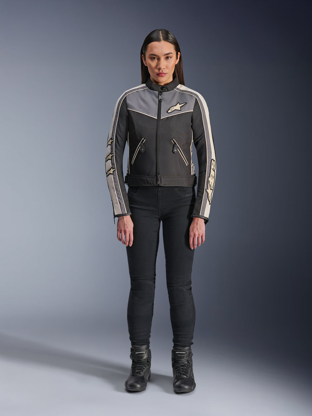 Femme portant la veste Alpinestars Stella T-Dyno WR, noir, gris foncé et écru, vue de face, veste de moto en textile au design d'inspiration vintage, avec logos sur la poitrine et les manches, portée avec un pantalon noir et des bottes
