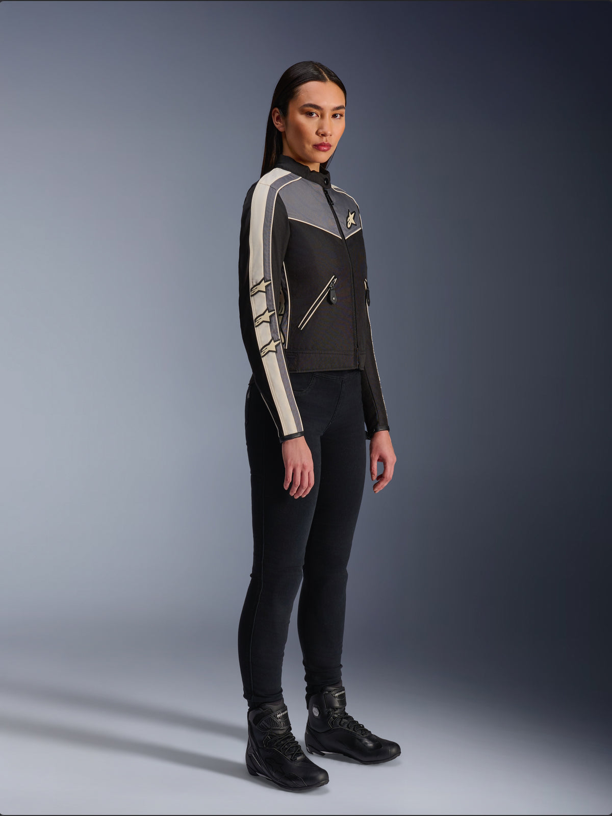 Veste Alpinestars Stella T-Dyno WR, veste de moto en textile pour femme, noir, gris foncé et écru, présentant un design d'inspiration vintage avec des bandes de manches crème et des logos étoiles, portée avec un pantalon noir et des bottes de moto, équipement de conduite urbaine d'inspiration héritage.