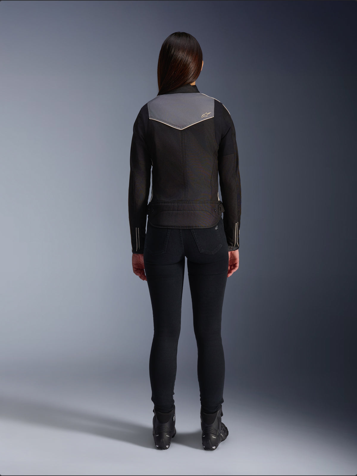 Vue arrière d'une motarde portant la veste Alpinestars Stella T-Dyno WR en Noir, Gris Foncé et Écru, accompagnée d'un pantalon slim noir et de bottes de moto noires, mettant en valeur le panneau supérieur du dos gris avec le logo Alpinestars et les détails de finition écru.