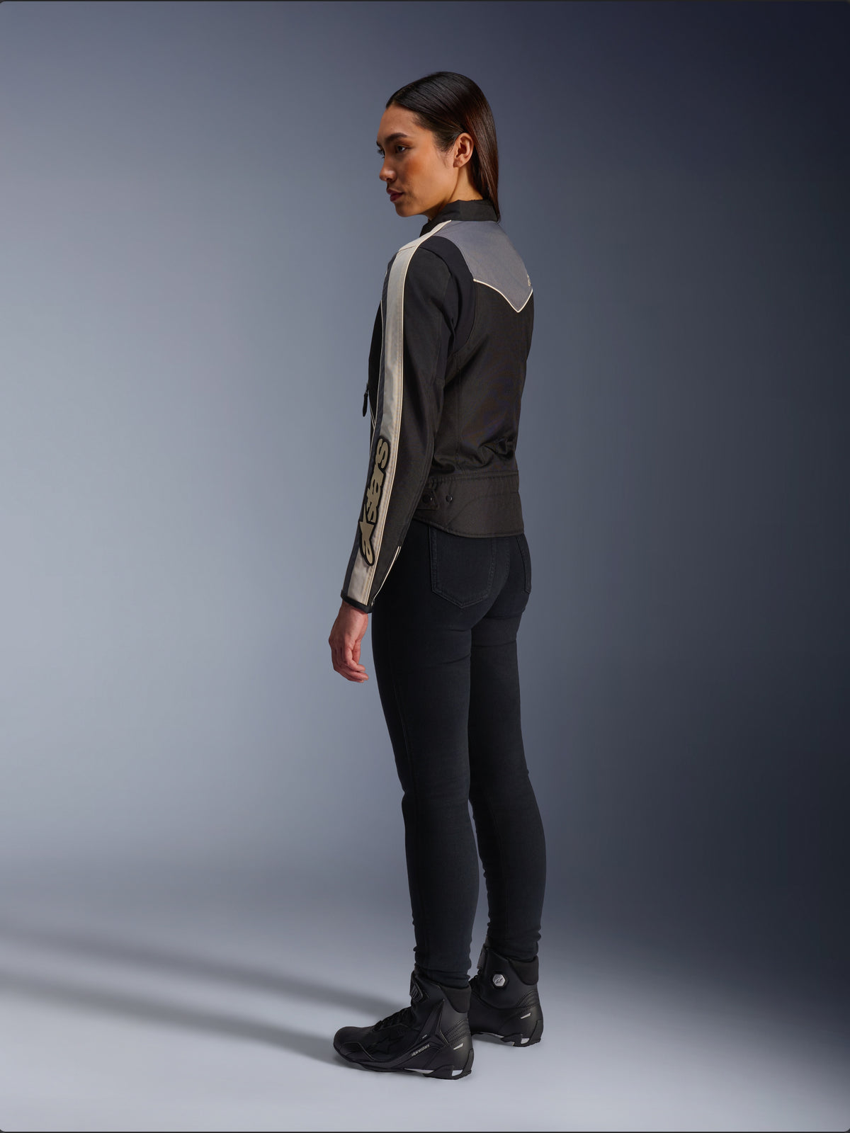 Mannequin femme vue de profil et de dos portant la veste Alpinestars Stella T-Dyno WR en Noir Gris Foncé Écru, au design d'inspiration vintage avec des bandes écrues sur les manches et un empiècement gris dans le dos, portée avec un pantalon noir et des chaussures de moto noires.
