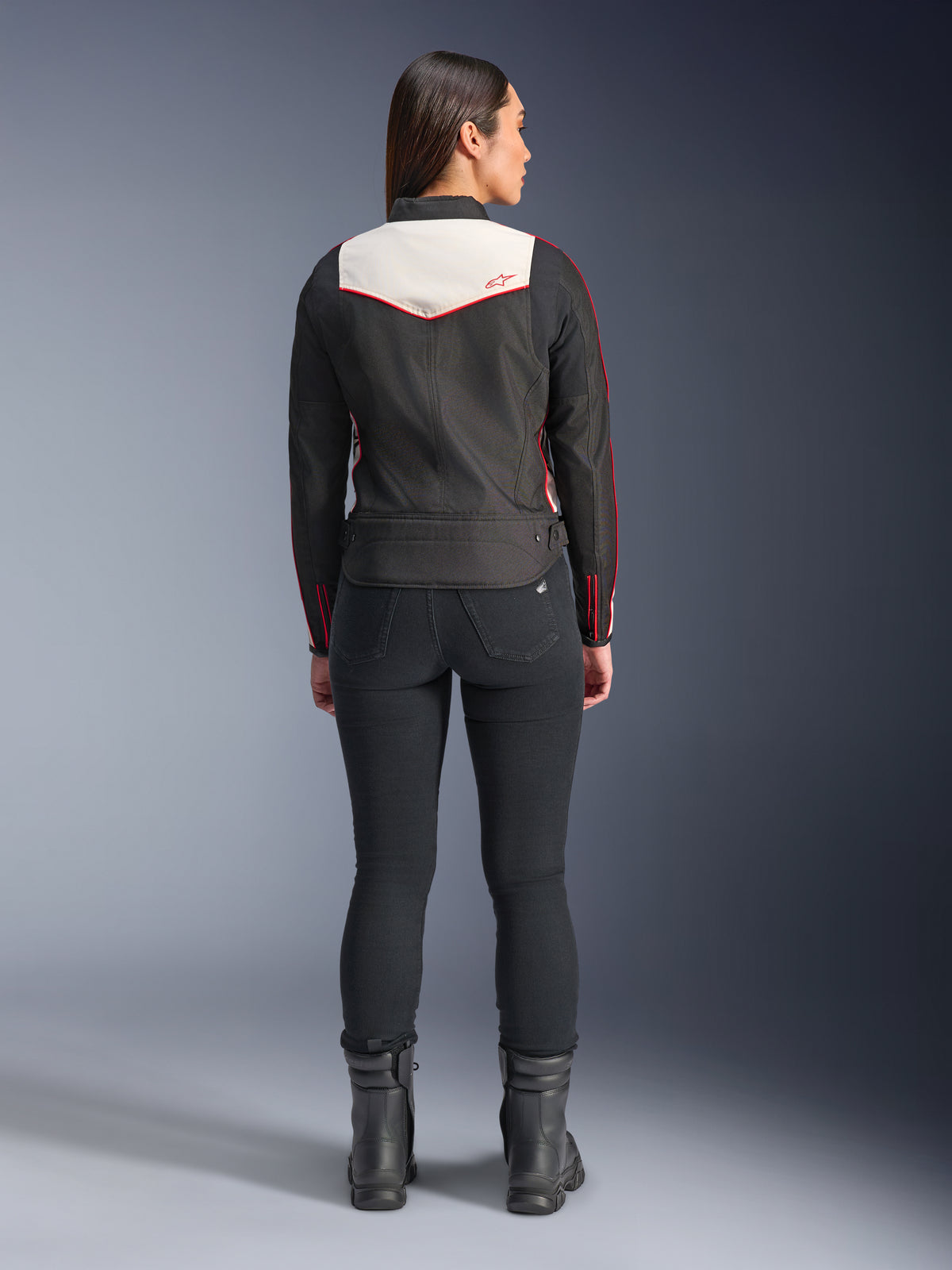 Vue arrière d'un mannequin femme portant la veste Alpinestars Stella T-Dyno WR en Noir Écru Rouge Éclatant, dotée d'un empiècement dorsal crème et de logos rouges, assortie d'un pantalon et de bottes noirs, une veste de moto en textile d'inspiration vintage.
