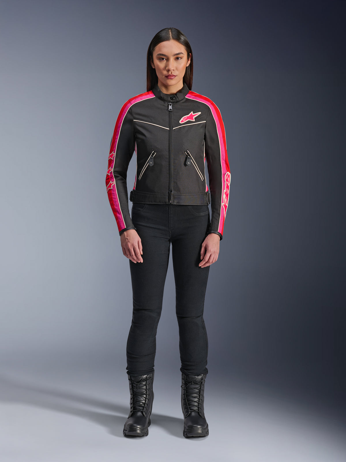 Vrouw die Alpinestars Stella T-Dyno WR jas draagt, zwart met helderrode en fuchsia accenten, vintage-stijl textiele jas met roze logo en mouwstrepen, gedragen met zwarte broek en veterlaarzen, urban motorkleding