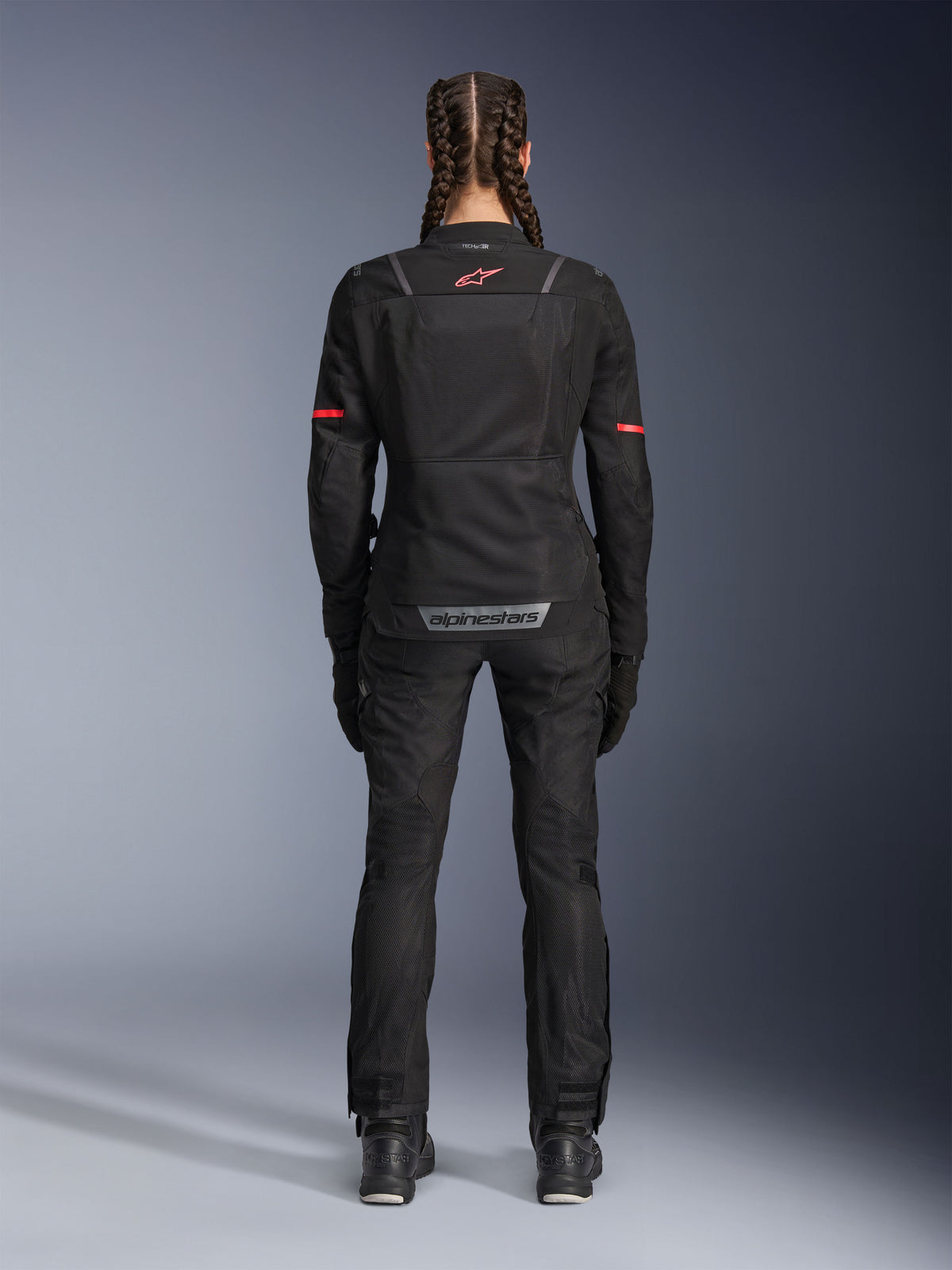 Alpinestars Stella ST-2 Air Jacke, Motorradjacke für Damen, Schwarz, Rückansicht mit rotem Logo auf dem oberen Rücken und grauem Branding am unteren Rücken, getragen mit passender schwarzer Motorradhose und Stiefeln, modernes Touring-Design mit Mesh-Einsätzen und reflektierenden Details.