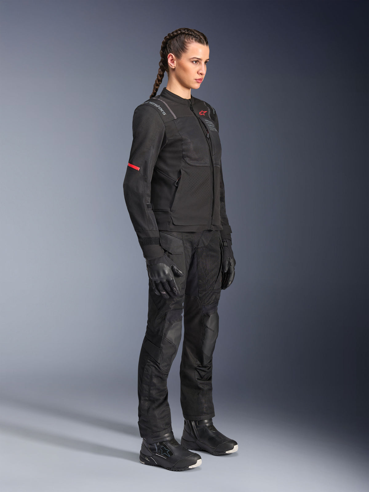 Weibliches Model trägt die Alpinestars Stella ST-2 Air Jacke in Schwarz, kombiniert mit passender schwarzer Motorradhose, Handschuhen und Stiefeln, mit Mesh-Belüftungseinsätzen und dezenten roten Details, elegantes modernes Touren-Outfit