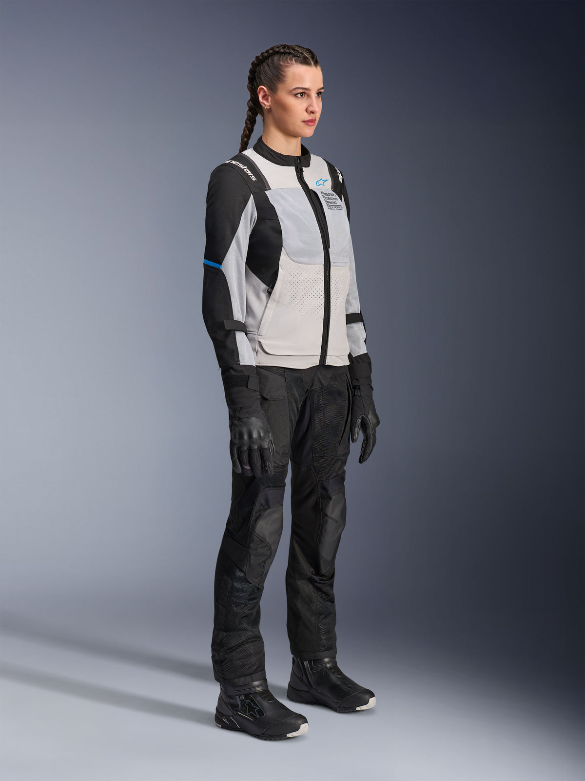 Model draagt de Alpinestars Stella ST-2 Air Jacket in Ice Gray en zwart, technische motorjas voor dames met meshpanelen en softshell-constructie, gecombineerd met een zwarte adventure touring-broek en laarzen, voorzien van blauwe accenten en een modern rij-ontwerp