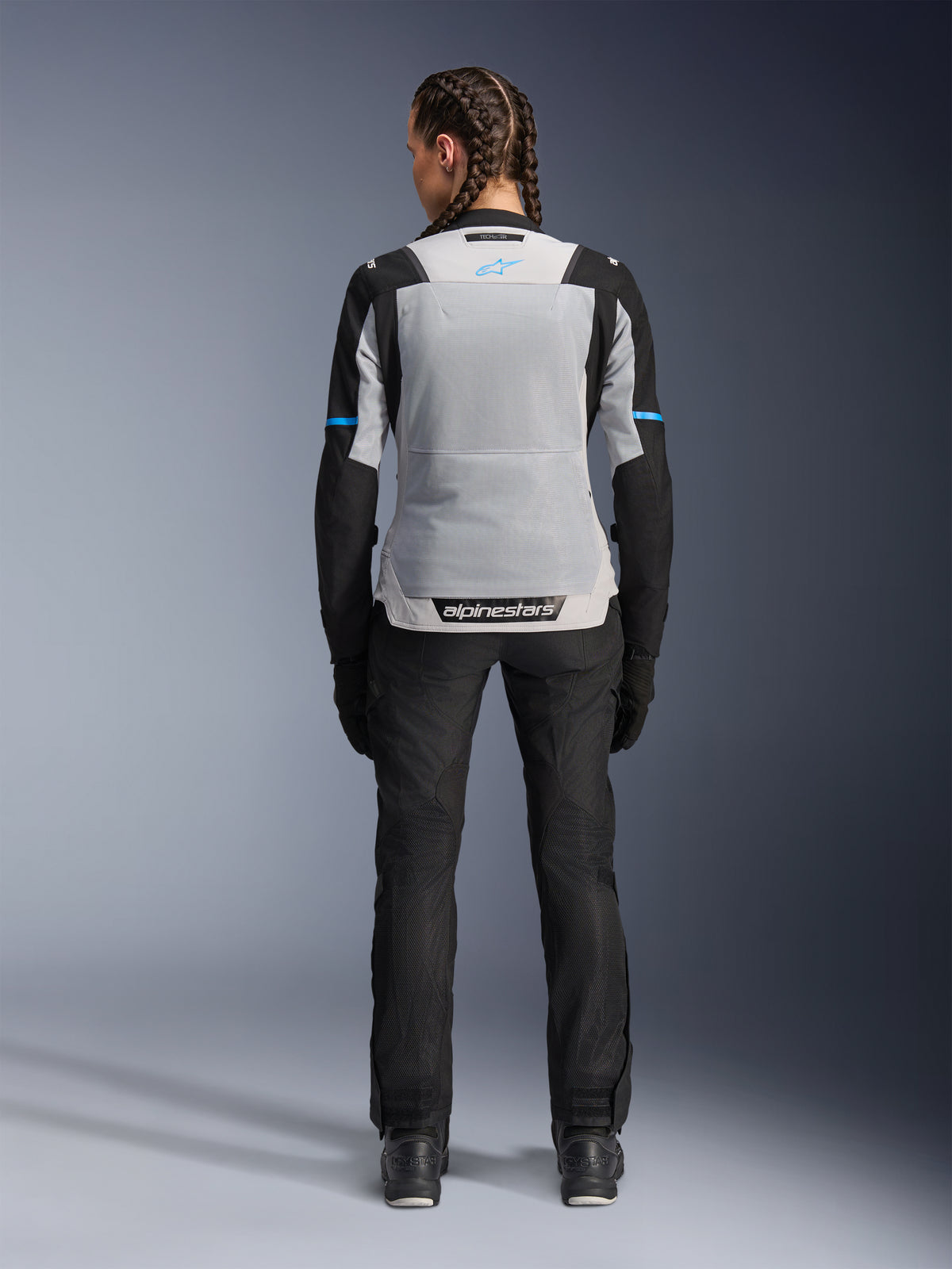 Alpinestars Stella ST-2 Air Jacket, Ice Gray Black, achteraanzicht op vrouwelijk model, voorzien van een groot lichtgrijs mesh rugpaneel, zwarte mouwen met blauwe streepaccenten, Alpinestars-branding op de onderrug, gecombineerd met zwarte technische motorbroek en laarzen, zomerse motoruitrusting
