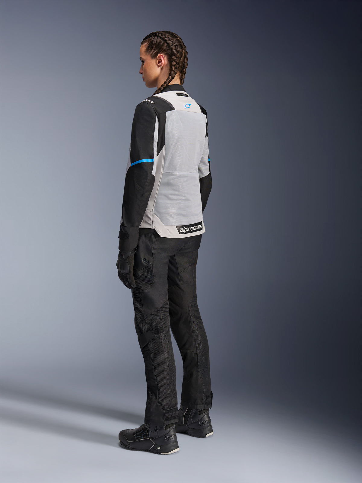 Vrouwelijke motorrijder draagt het Alpinestars Stella ST-2 Air Jacket in Ice Gray en zwart, driekwart achteraanzicht met focus op het grote lichtgrijze mesh rugpaneel, zwarte mouwen met een blauwe accentstreep en een blauw Alpinestars-logo, gecombineerd met een zwarte motorbroek, handschoenen en laarzen voor een complete adventure touring-outfit