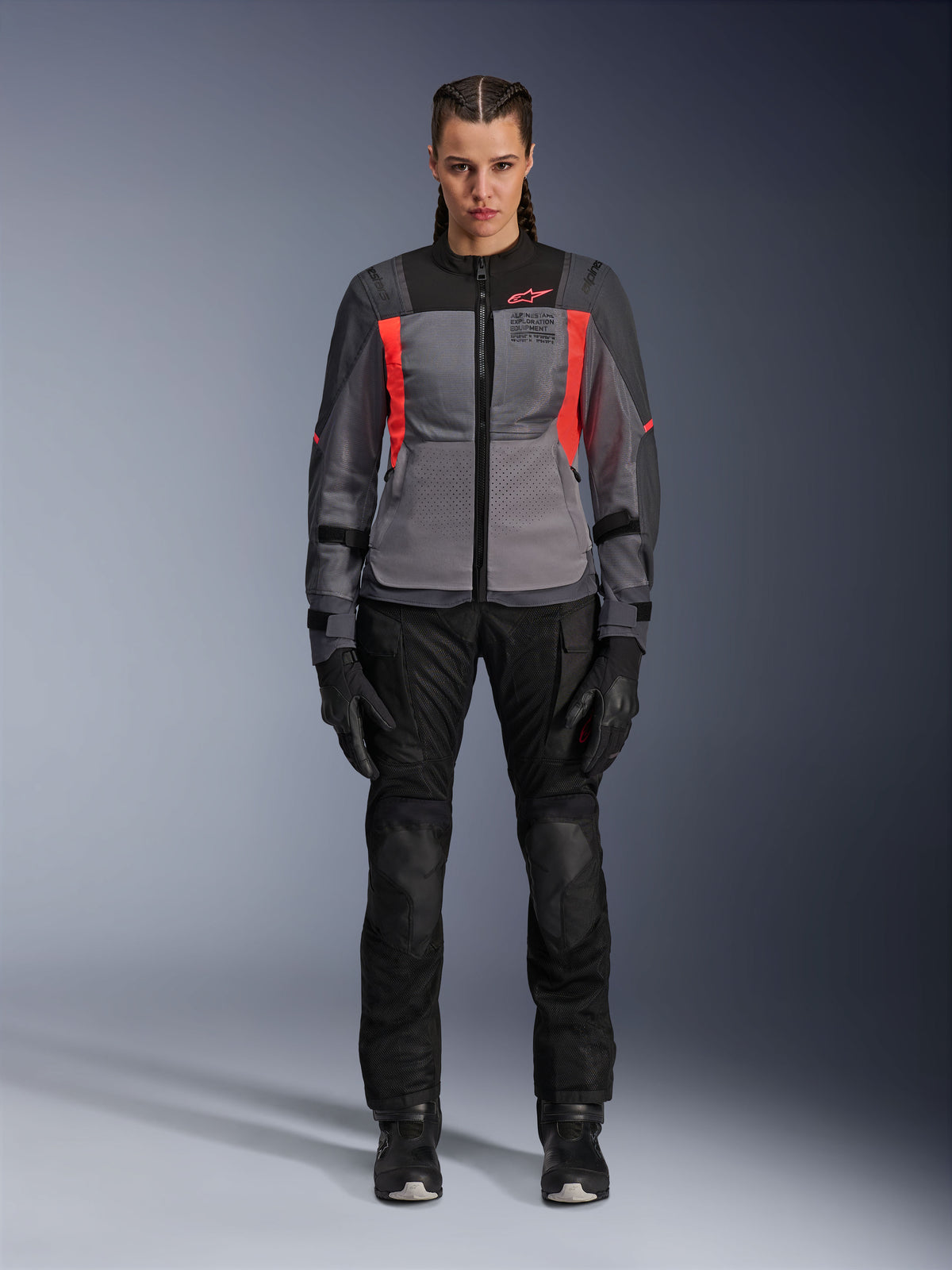 Vrouw die de Alpinestars Stella ST-2 Air Jacket draagt in Deep Gray, Dark Gray en Fluo Coral, gecombineerd met een zwarte motorbroek en handschoenen, vooraanzicht, moderne mesh adventure touring-uitrusting voor dames