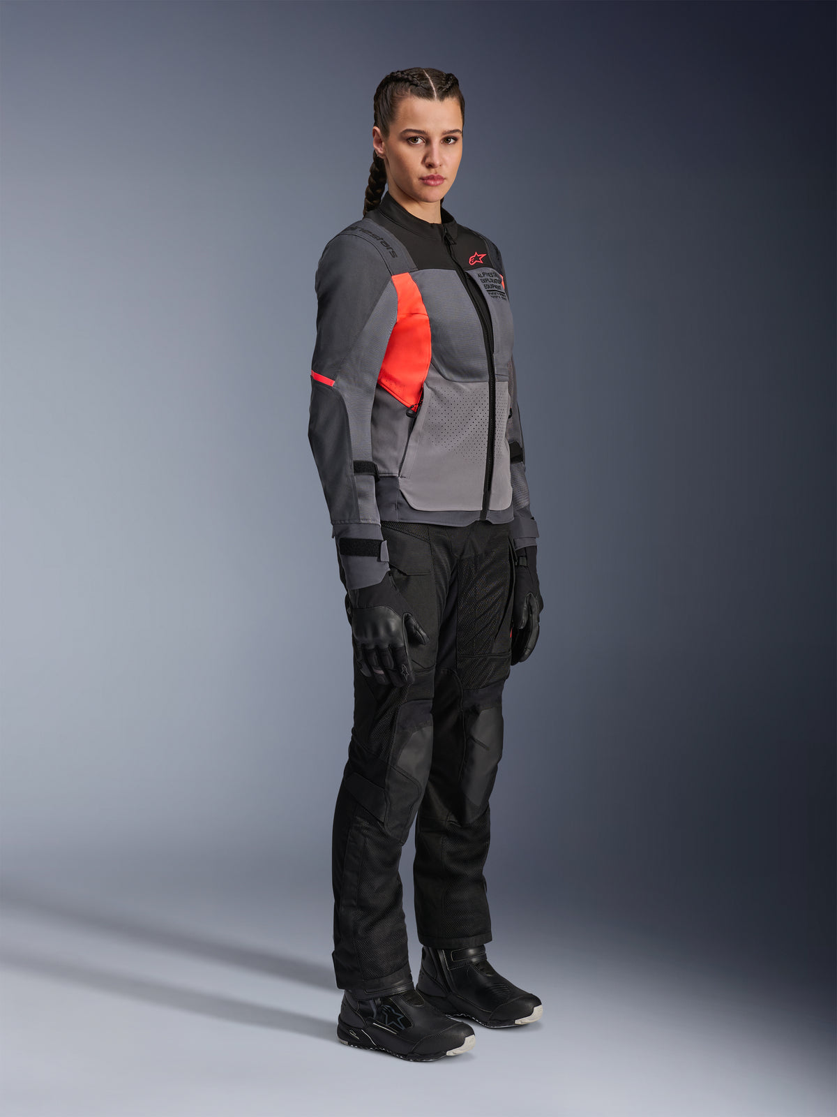 Vrouwelijke motorrijder die de Alpinestars Stella ST-2 Air Jacket draagt in Deep Gray, Dark Gray en Fluo Coral, gecombineerd met een zwarte motorbroek, handschoenen en laarzen, voorzien van een modern touring-ontwerp met mesh-panelen en reflecterende details.