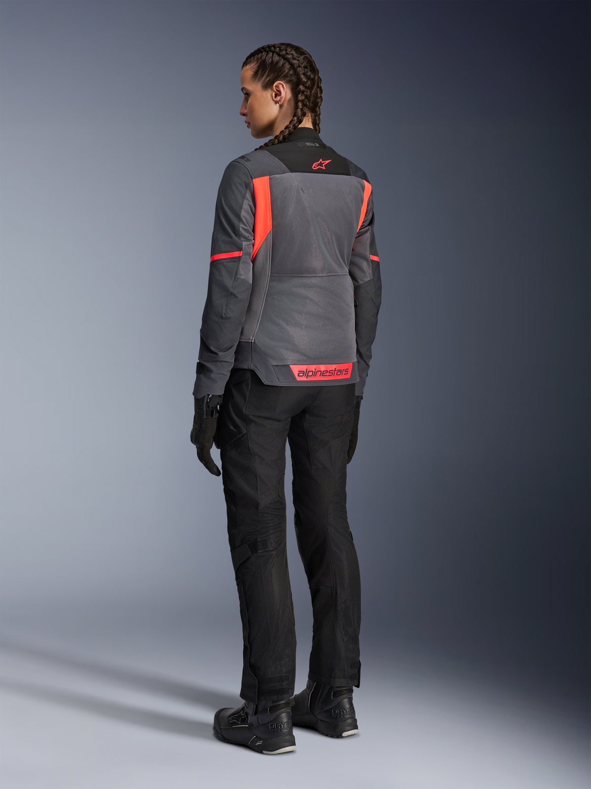 Vrouw draagt de Alpinestars Stella St-2 Air Jas, motor-toerjas, Deep Gray en Dark Gray met Fluo Coral accenten, gecombineerd met een zwarte motorbroek en handschoenen, achteraanzicht met grote mesh-panelen en reflecterende branding in koraalrood, ontworpen voor zomerse motoravonturen