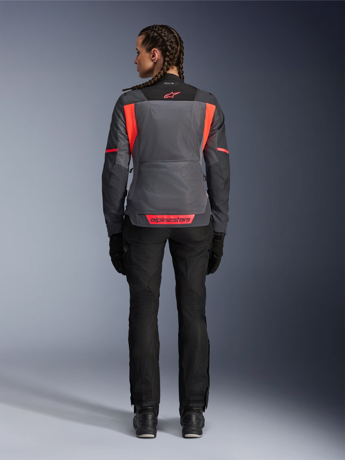 Achteraanzicht van een model dat de Alpinestars Stella St-2 Air damesjas draagt in Deep Gray en Dark Gray met Fluo Coral accenten, voorzien van grote mesh ventilatiepanelen op de rug en reflecterende logodetails, gecombineerd met een zwarte motorbroek en handschoenen.