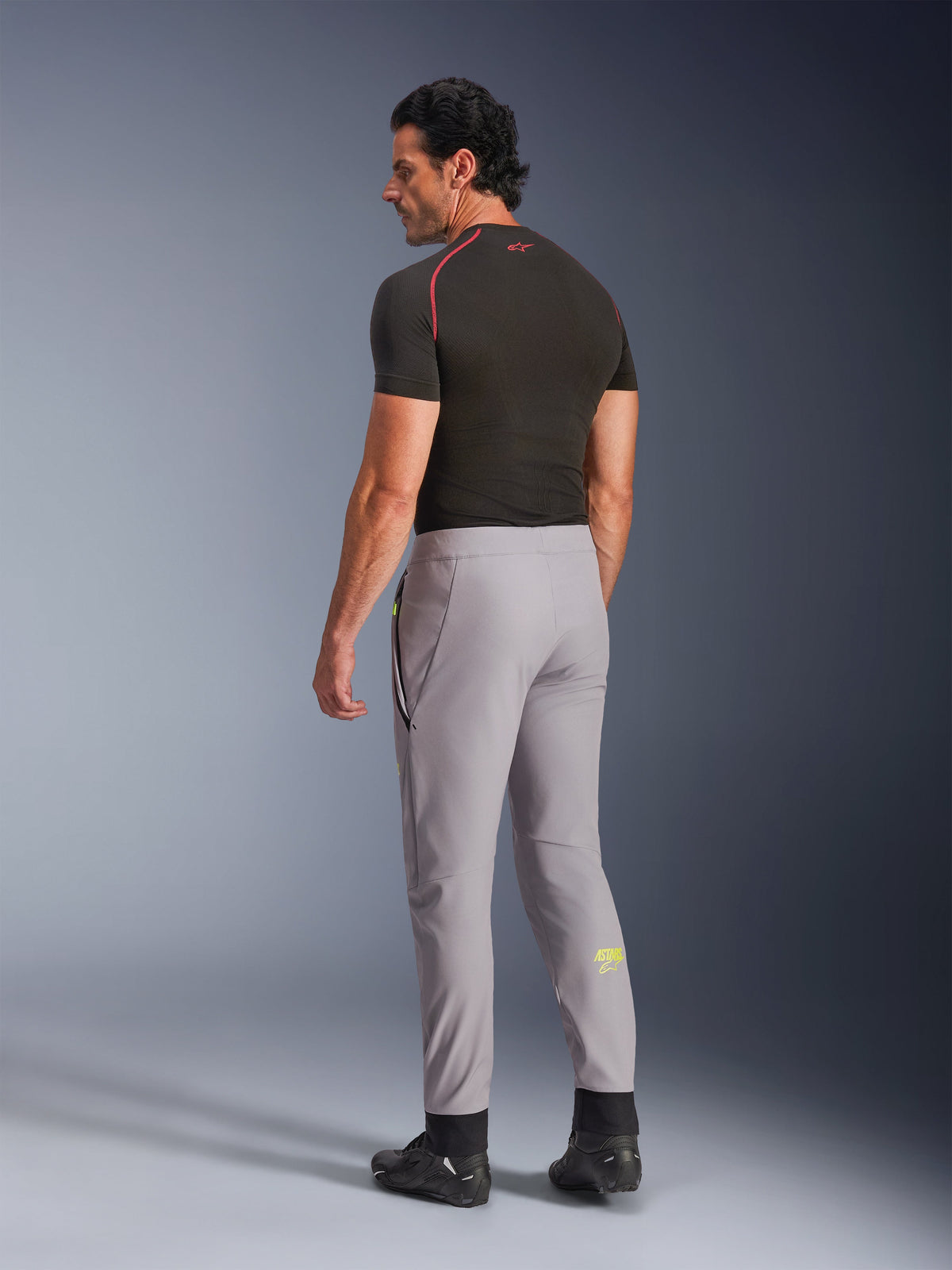 Alpinestars Aeroshell broek, Mid Gray Yellow Fluo, achteraanzicht van een mannelijk model dat een stretch softshell motorbroek draagt, voorzien van een geel Astars-logo op de linkerkuit, gecombineerd met een zwarte basislaag en motorschoenen voor een modern sportief silhouet.