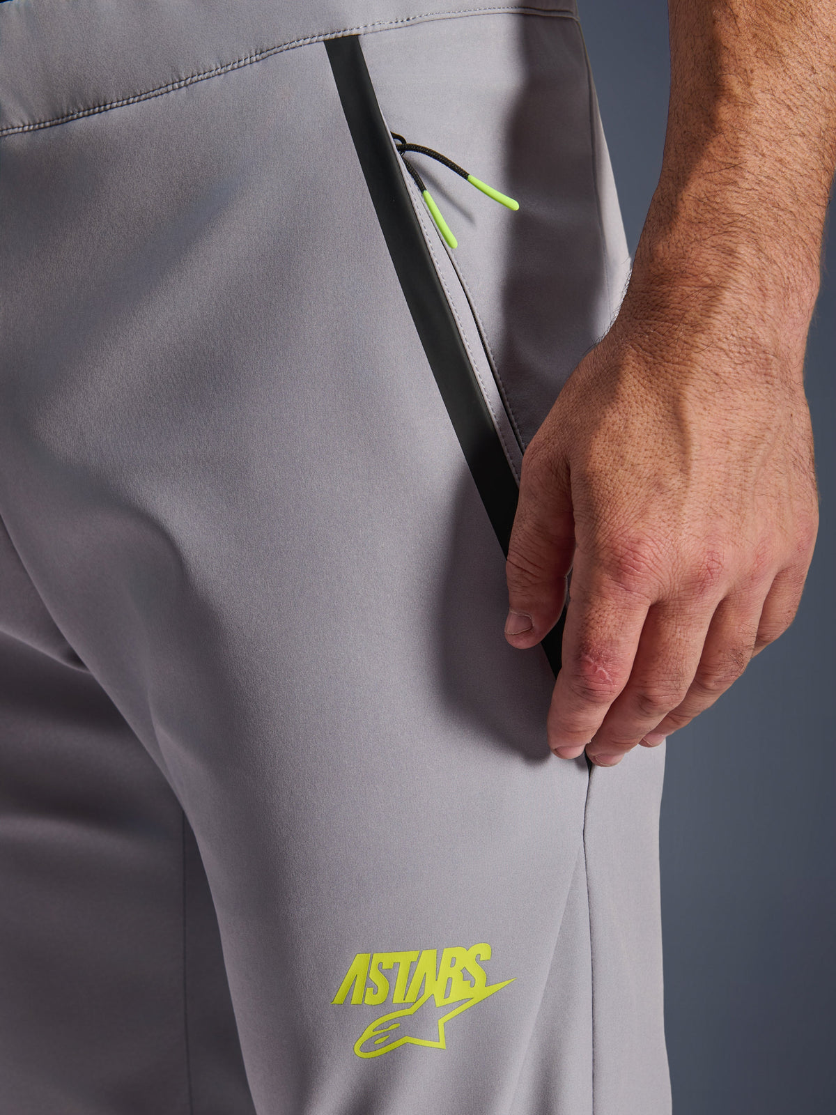 Alpinestars Aeroshell broek, middengrijs met fluorgele accenten, detailopname van de voorzak met rits met zwarte getapete bies en felgele trekkoordjes, geel ASTARS-logo op de dij, softshell stretchmateriaal voor motorrijden