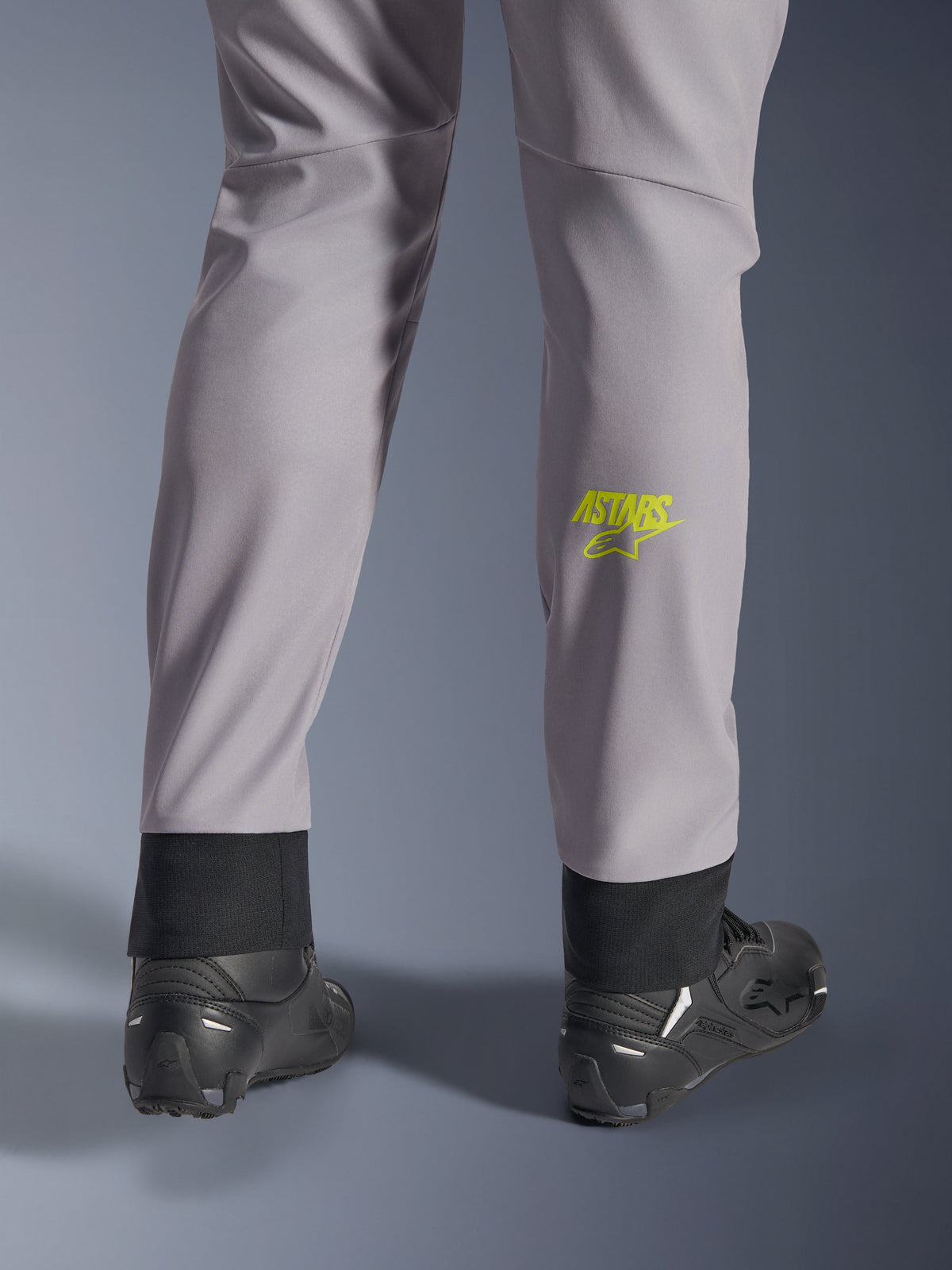 Alpinestars Aeroshell Pants, middengrijs met geel fluo Astars-logo op de kuit, van achteren gezien gedragen met zwarte motorschoenen, voorzien van een elastische onderzoom en een modern taps toelopend silhouet