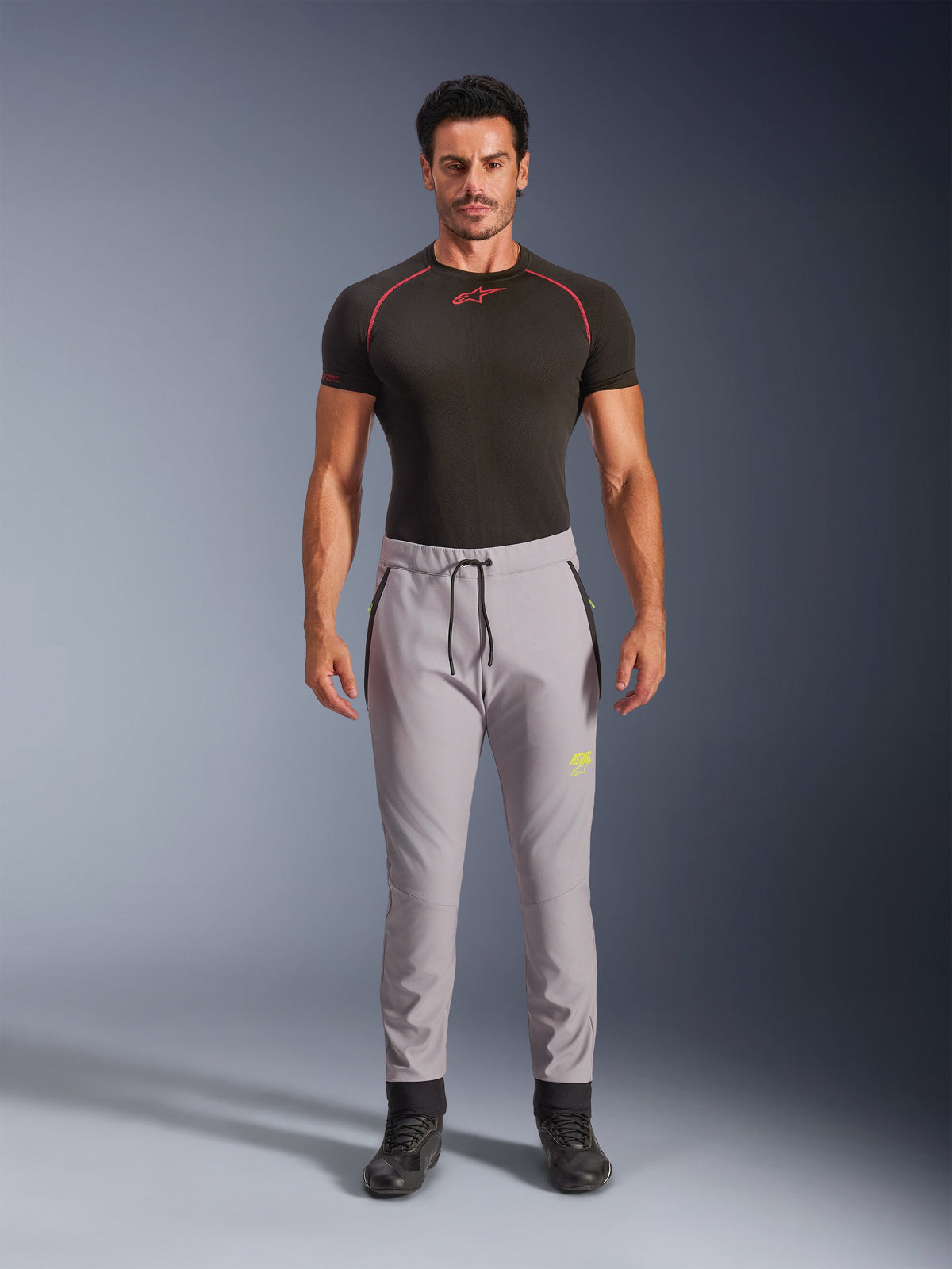 Alpinestars Aeroshell Pants in Mid Gray en Yellow Fluo, crossover sportieve softshellbroek met elastische tailleband en trekkoord, zakken met rits, mannelijk model draagt een zwart technisch shirt met korte mouwen, modern atletisch ontwerp voor road en casual gebruik