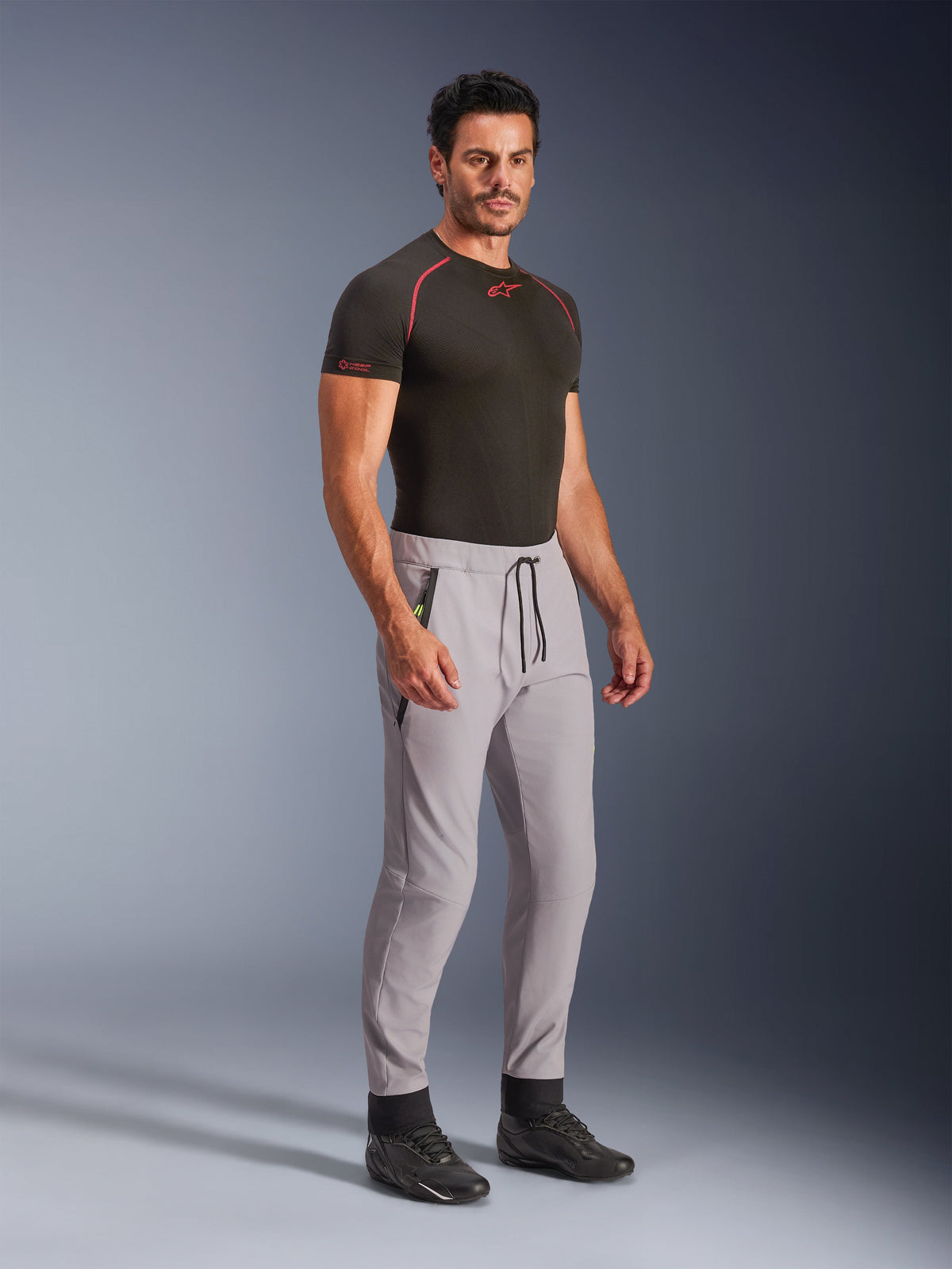 Alpinestars Aeroshell Pants, mid-grijze crossover softshell-broek met Yellow Fluo accenten, gedragen door een model met een zwarte basislaag en zwarte schoenen, voorzien van een moderne toelopende pasvorm, elastische tailleband met zwart trekkoord en zakken met ritssluiting.