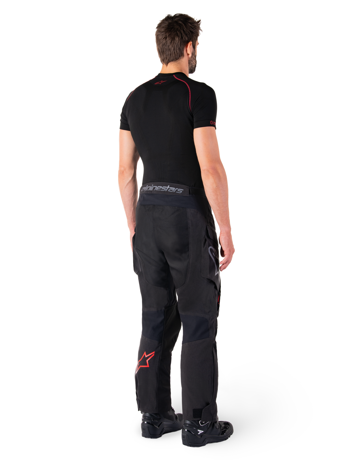 Alpinestars Hyde XT Drystar® broek, adventure touring broek, zwart en felrood, achteraanzicht van een rijder die de broek draagt met een zwarte basislaag en zwarte laarzen, met een duurzame textielconstructie, een rood sterlogo op het onderbeen en grijze Alpinestars-branding op de tailleband