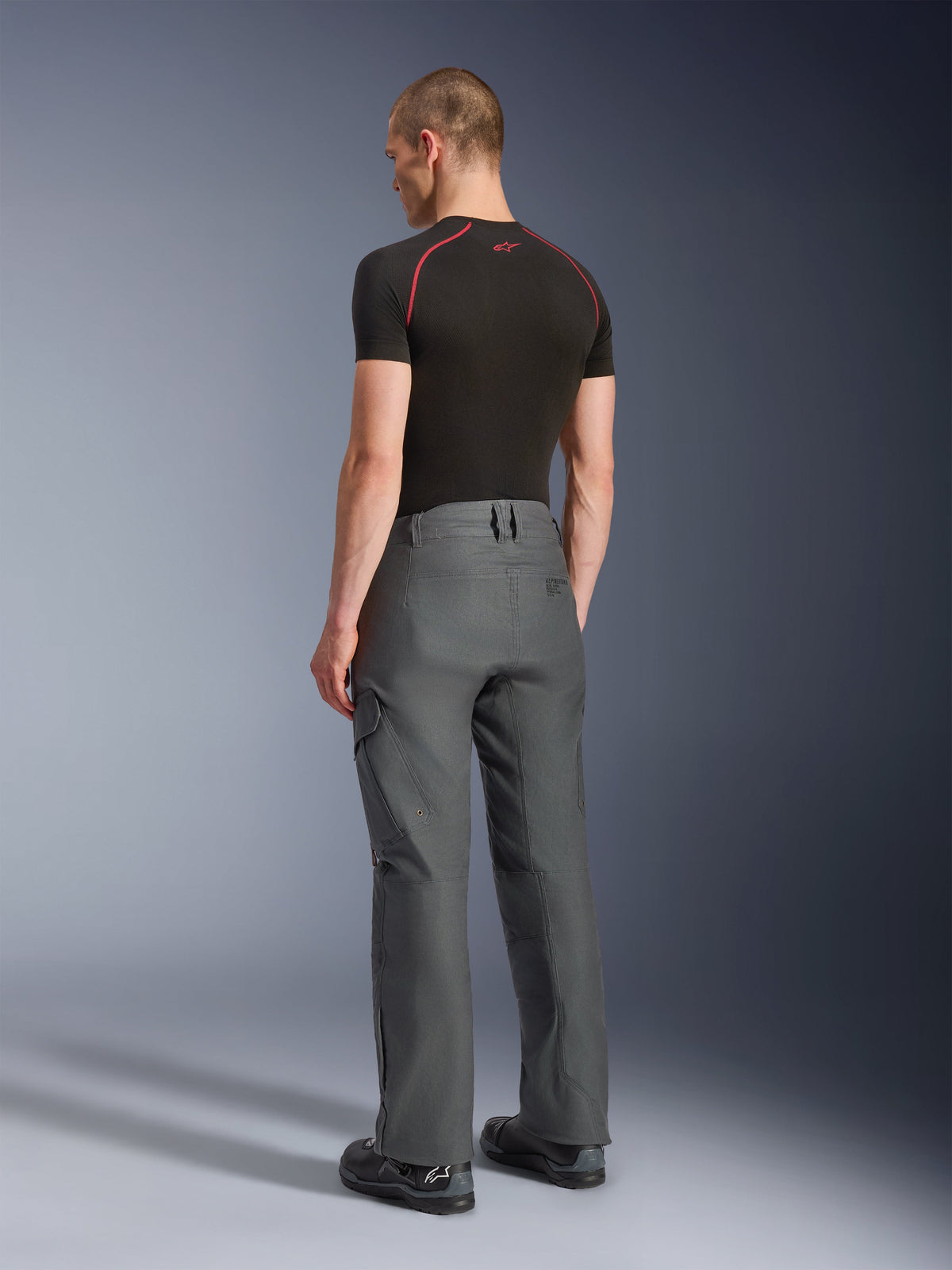 Pantalon Alpinestars Borrego Drystar®, pantalon de moto d'aventure, gris anthracite, vue arrière, aspect toile cirée avec poches cargo, associé à un t-shirt noir et des bottes de moto, design robuste pour la route et le touring d'aventure