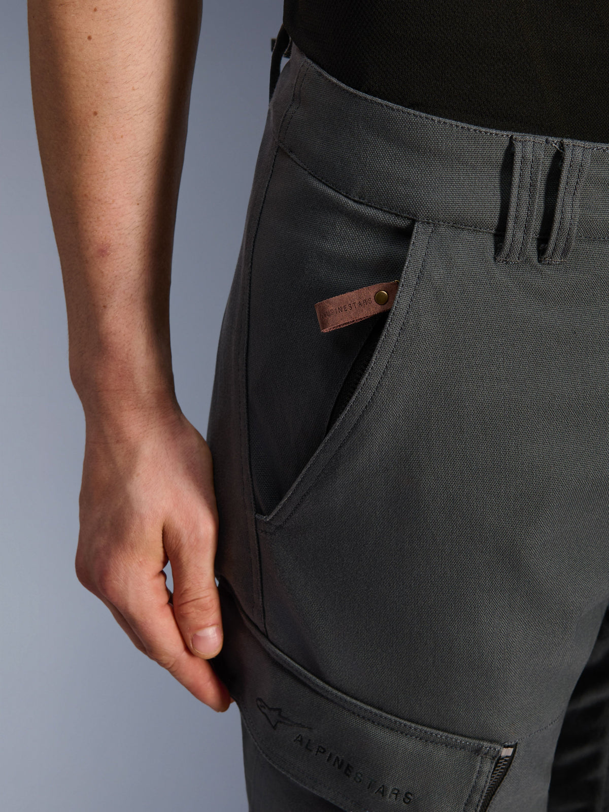 Pantalon Alpinestars Borrego Drystar®, pantalon de moto d'aventure, gris anthracite, gros plan sur le détail de la poche et de la hanche, texture en toile cirée avec une étiquette logo en cuir marron et logo Alpinestars brodé sur la poche cargo de la cuisse.